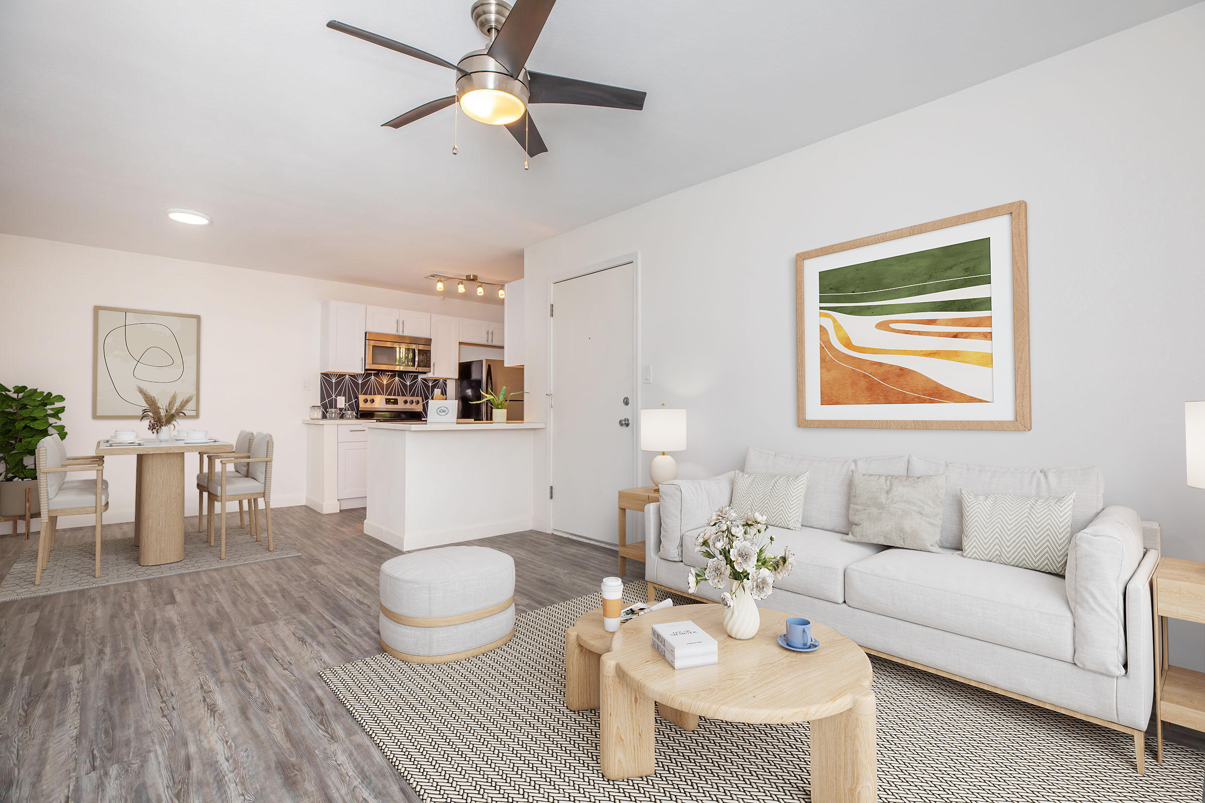 A modern, cozy living room featuring a light-colored sofa with decorative pillows, a wooden coffee table, and a round ottoman. The space includes a dining area with a table and chairs, and the kitchen is visible in the background. A large framed artwork adorns the wall, and a ceiling fan hangs above.