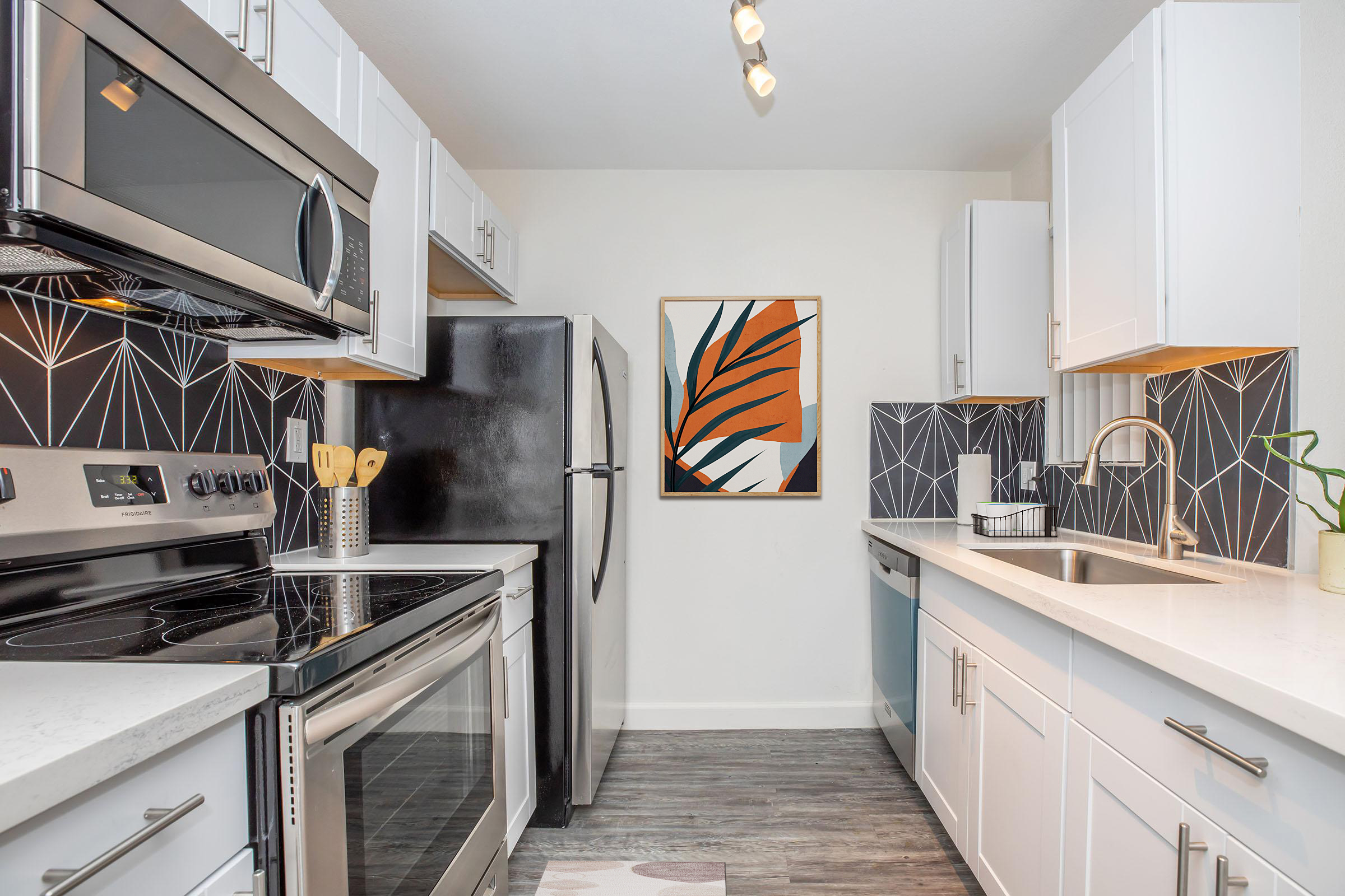 Modern kitchen featuring stainless steel appliances, including a microwave, oven, and refrigerator. The cabinets are white, complemented by a geometric black and white backsplash. A framed artwork with abstract leaves hangs on the wall, and there is a small plant near the sink. Bright lighting enhances the space.