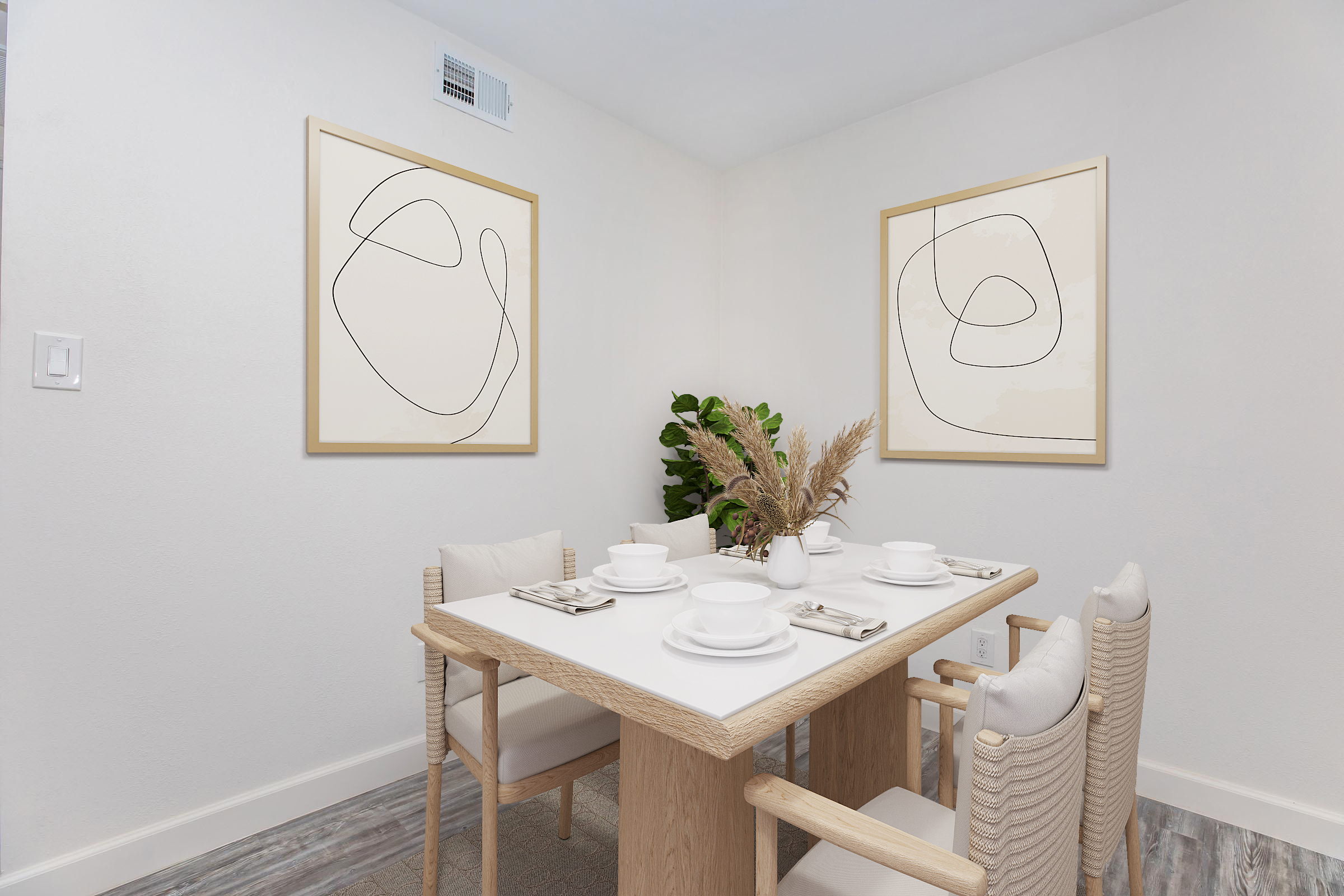 A cozy dining area featuring a white table set with dishes, cutlery, and a decorative vase of dried plants. Two abstract line art pieces are framed on the walls, and a green plant adds a touch of nature to the serene, minimalist decor. The space has light-colored walls and a neutral aesthetic.