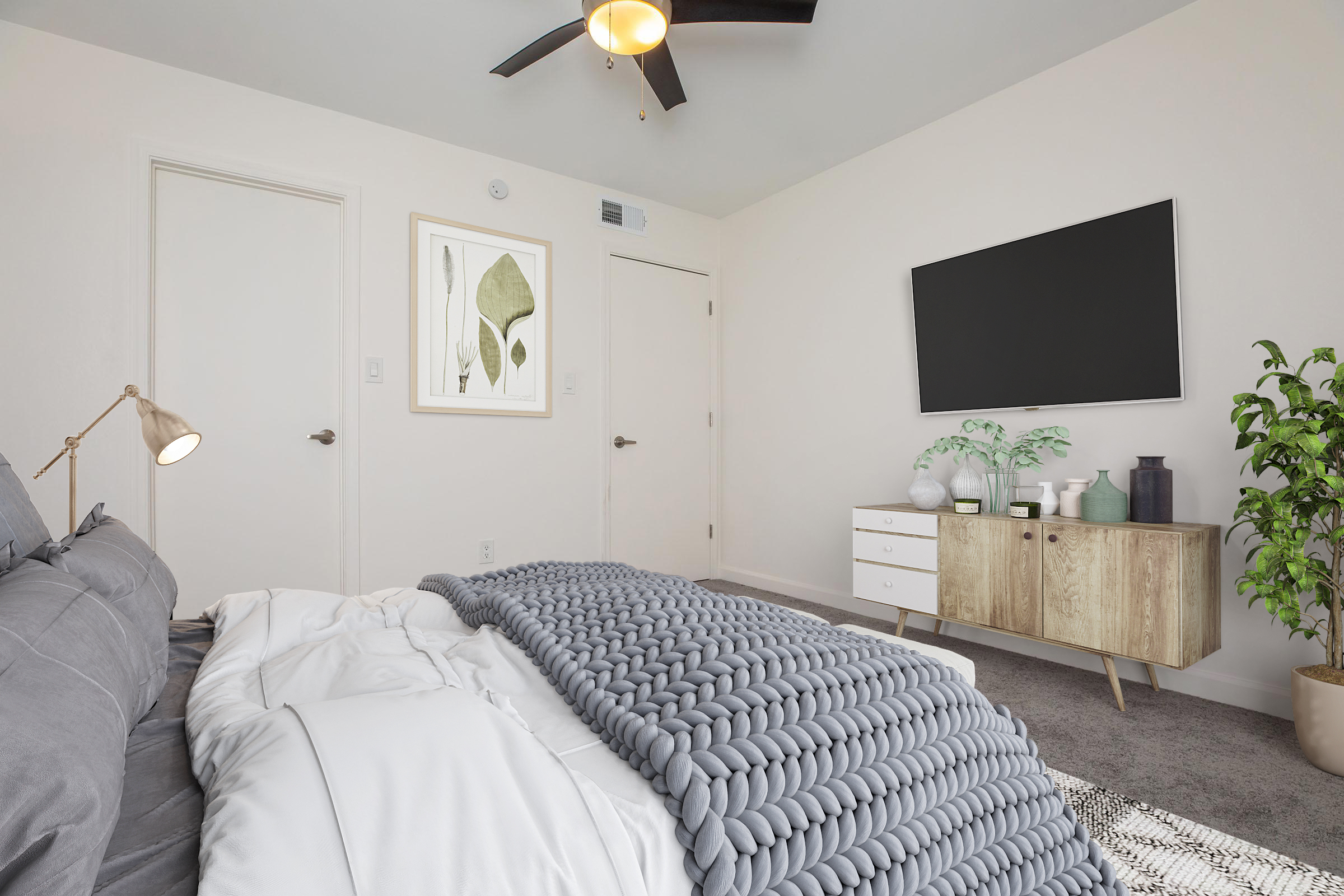 A modern bedroom featuring a cozy bed with a chunky knit blanket, a ceiling fan, a minimalist dresser with decorative vases, and a flat-screen TV mounted on the wall. The room is softly lit, with a piece of framed artwork depicting leaves and a potted plant in the corner.