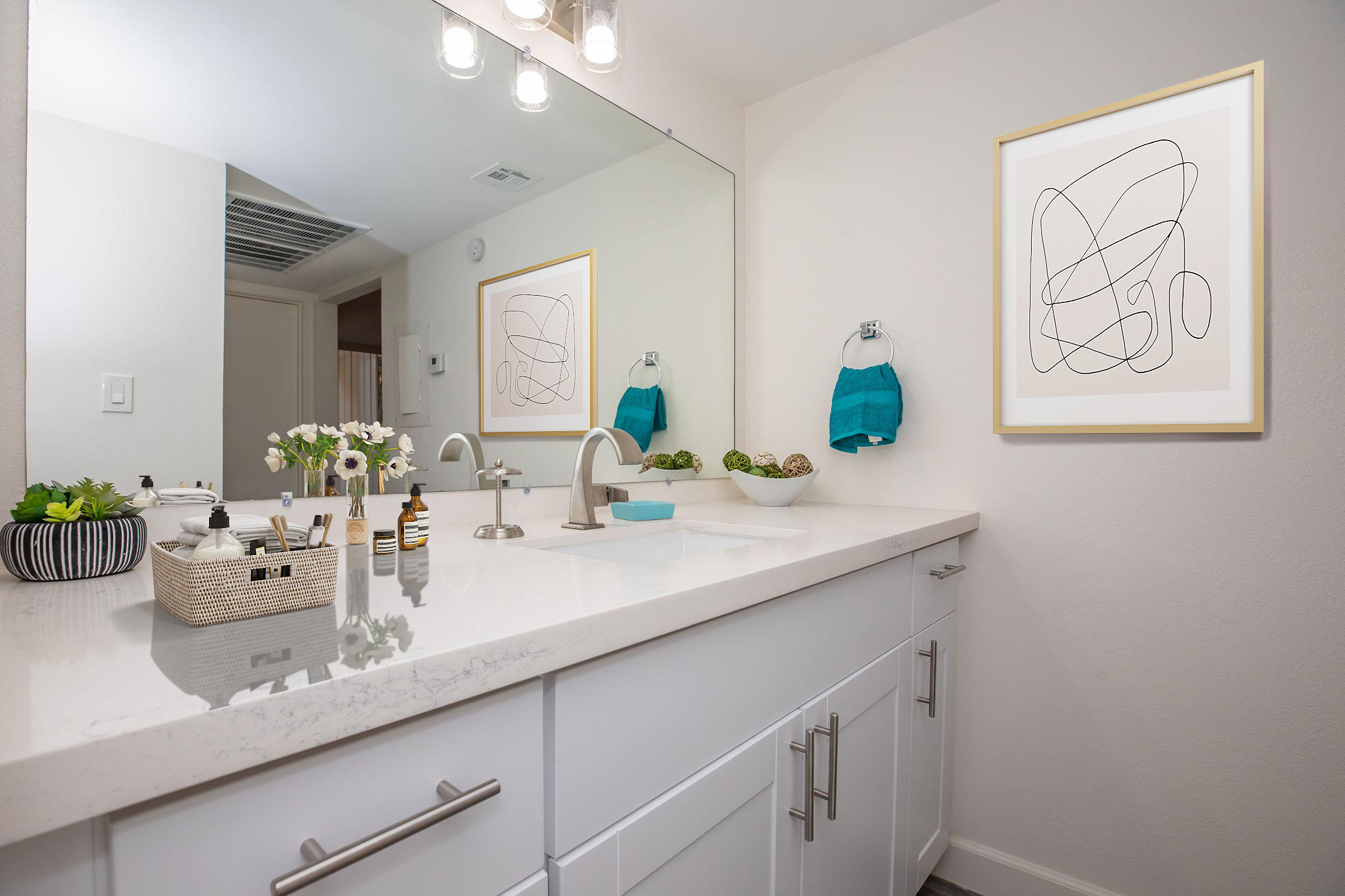 A modern bathroom featuring a white countertop with various toiletries, a decorative basket, and potted plants. Above the sink is a large mirror and artwork showcasing abstract lines. Two towels hang on a bar, and a light fixture illuminates the space, creating a fresh and stylish atmosphere.