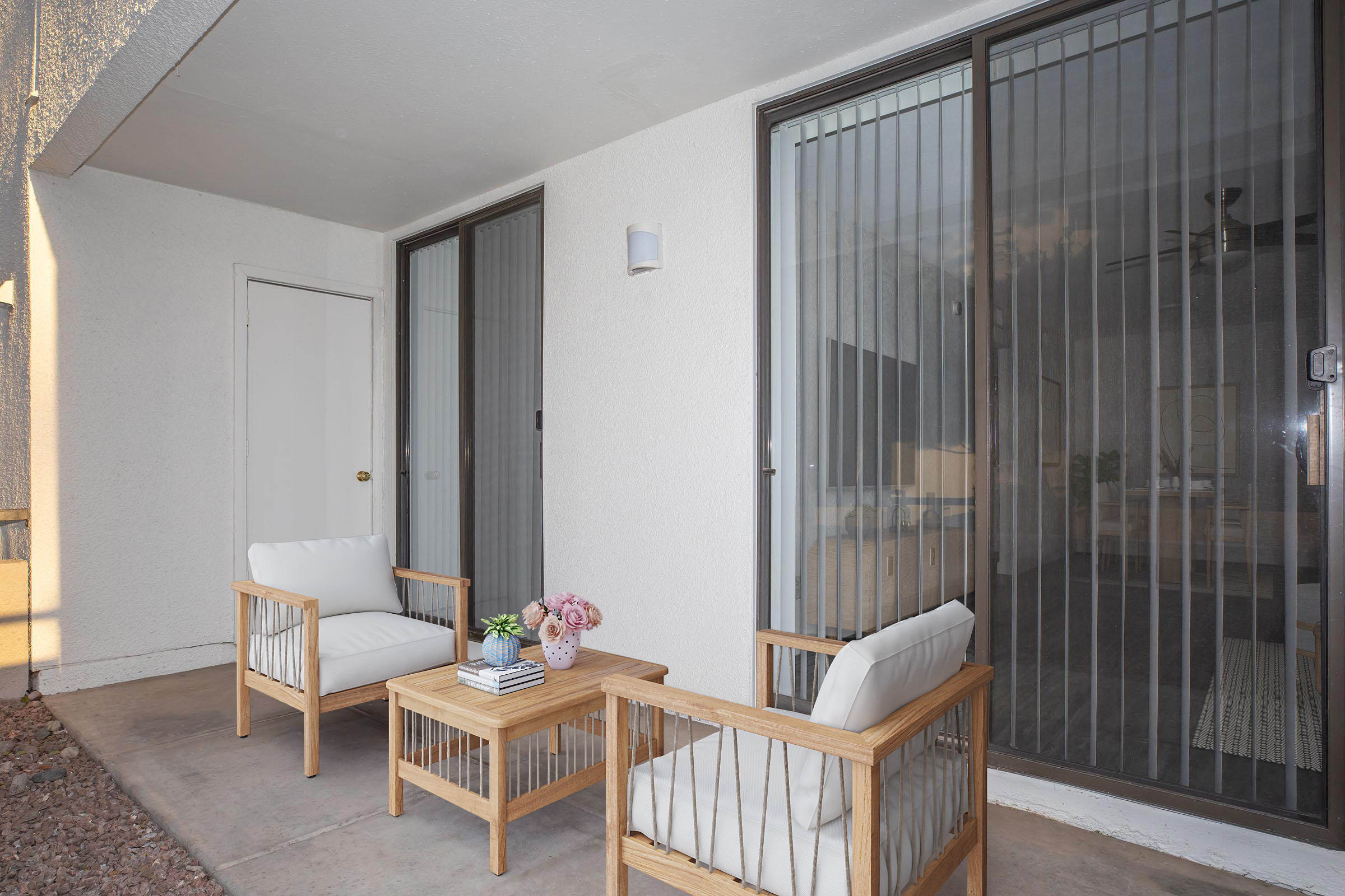 A small outdoor patio featuring two wooden chairs with white cushions and a small wooden table. A vase with flowers is on the table. The space has large glass sliding doors with vertical bars and a light-colored wall. The setting is cozy and inviting, perfect for relaxation.