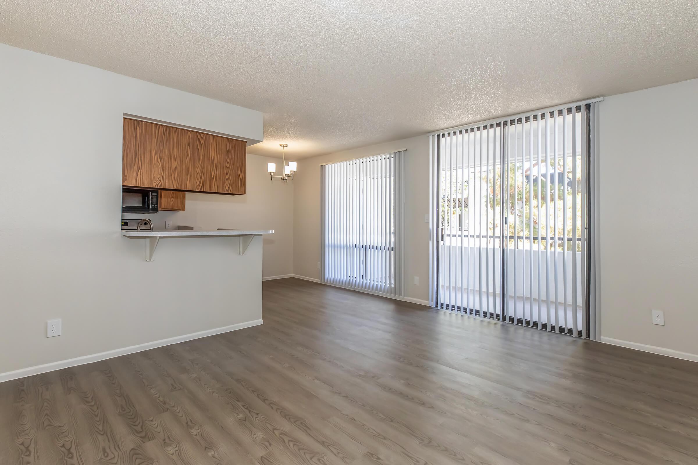 Spacious living area with light wood flooring, featuring large windows with vertical blinds, a dining area with a chandelier, and an open kitchen with wooden cabinetry and a microwave. The walls are painted in a soft neutral color, creating a bright and inviting atmosphere.