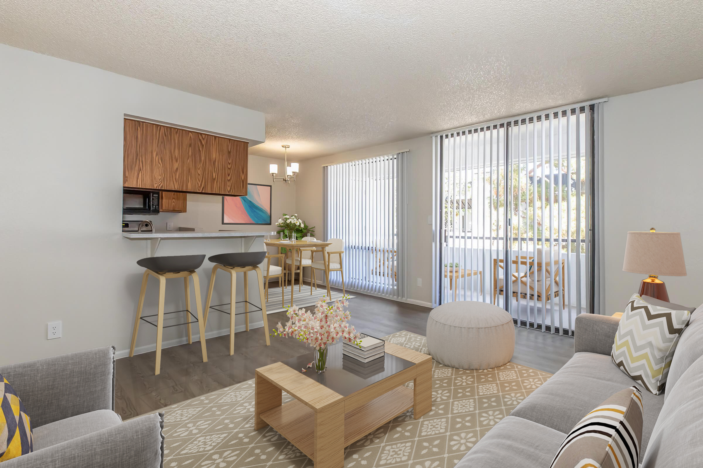 A modern living room featuring a light gray sofa with patterned cushions, a coffee table adorned with flowers, and a cozy area rug. The kitchen area has wooden cabinets, bar stools, and a small dining table with chairs. Large windows with vertical blinds provide natural light, opening to a patio.