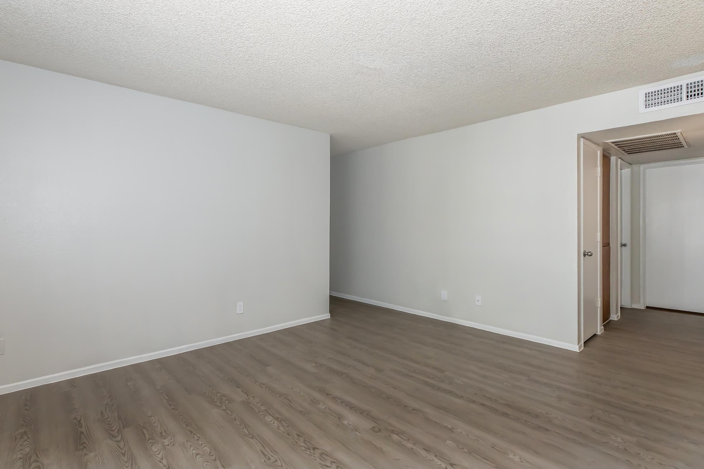 An empty room with light-colored walls and a textured ceiling. The flooring is a light wood laminate. There are two doors visible: one leading to a closet and another to an adjoining space. The room has a spacious, uncluttered look with natural light.