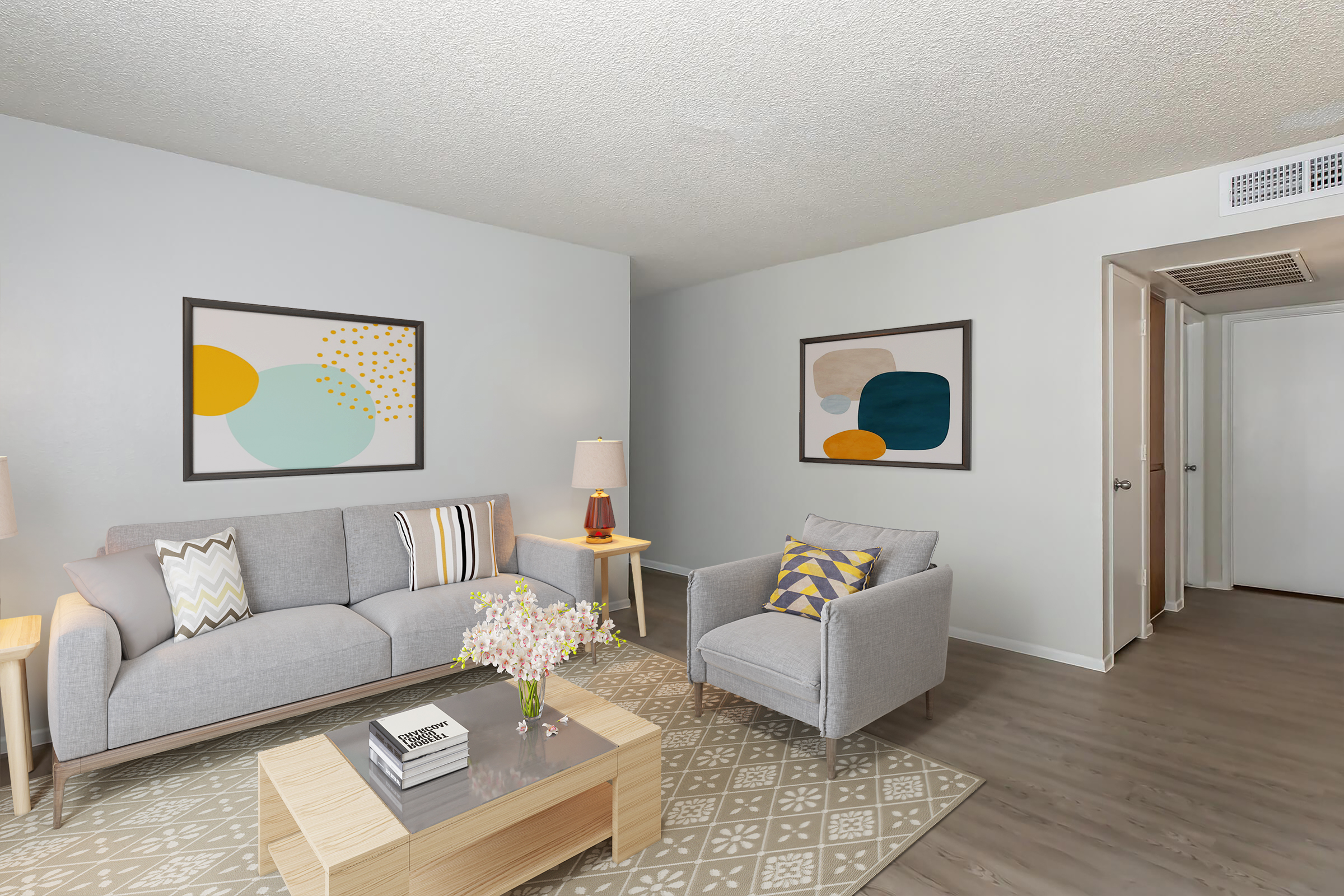 A cozy living room featuring a gray sofa with patterned cushions, an accent chair, a wooden coffee table with a decorative book, and a vase of flowers. The walls are adorned with abstract art pieces in soft colors, and the space has light-colored flooring and neutral walls, creating a welcoming atmosphere.