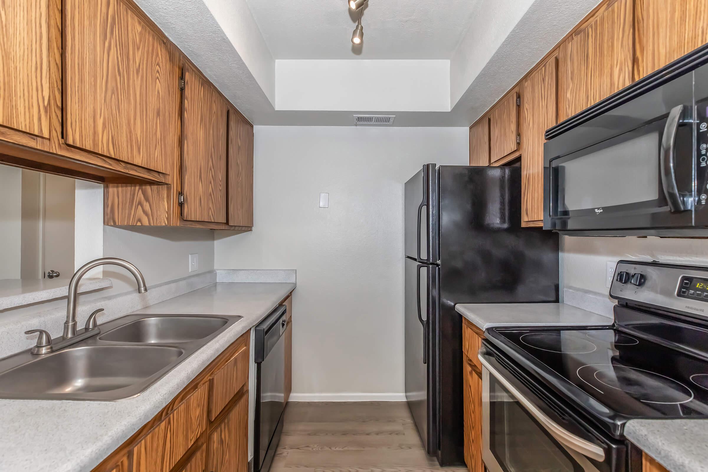 A modern kitchen featuring wooden cabinets, stainless steel appliances, and a double sink. The kitchen includes an electric stove, dishwasher, and refrigerator, with a light-colored countertop and neutral walls, providing a bright and functional space.