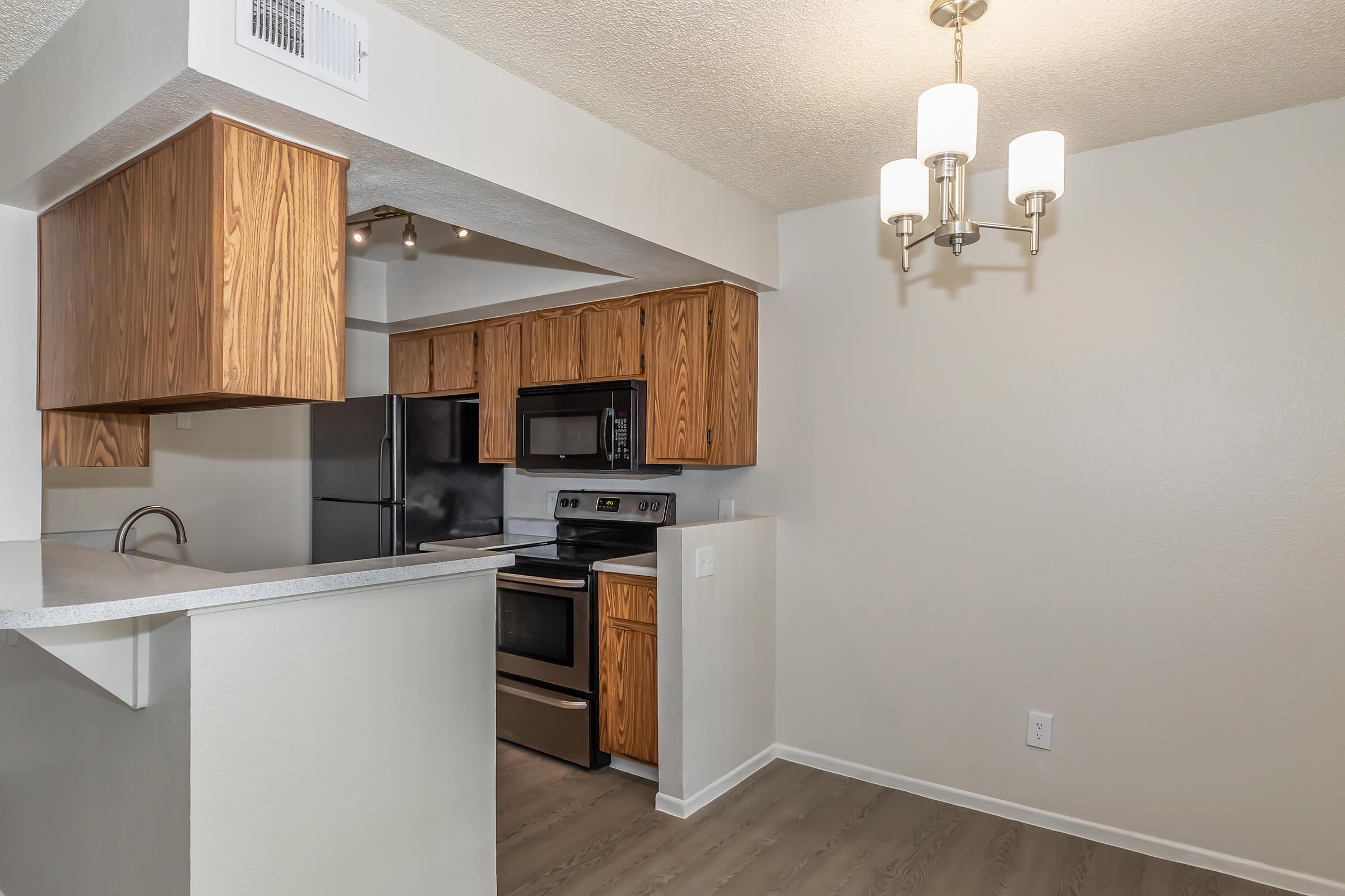 A modern kitchen with wooden cabinets and stainless steel appliances, including a stove, microwave, and refrigerator. The countertop has a minimalist design, and there's a chandelier above the dining area. The walls are light-colored, and the flooring is a contemporary laminate.