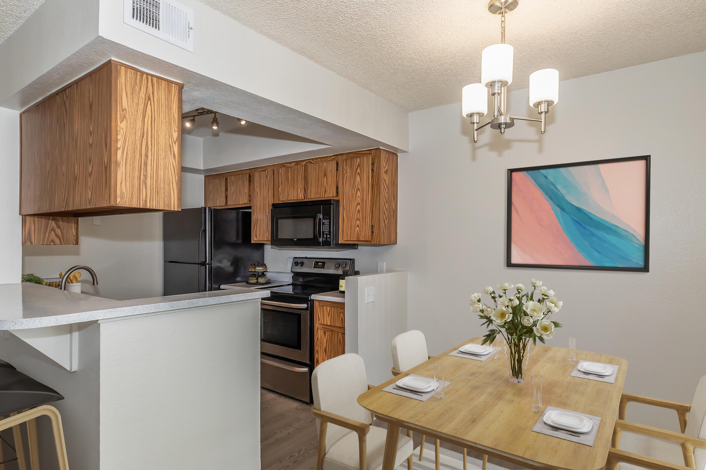 A modern kitchen with wooden cabinets, stainless steel appliances, and a dining area featuring a wooden table set for four. A bouquet of white flowers decorates the table, and a large abstract painting hangs on the wall, complementing the light and airy atmosphere.