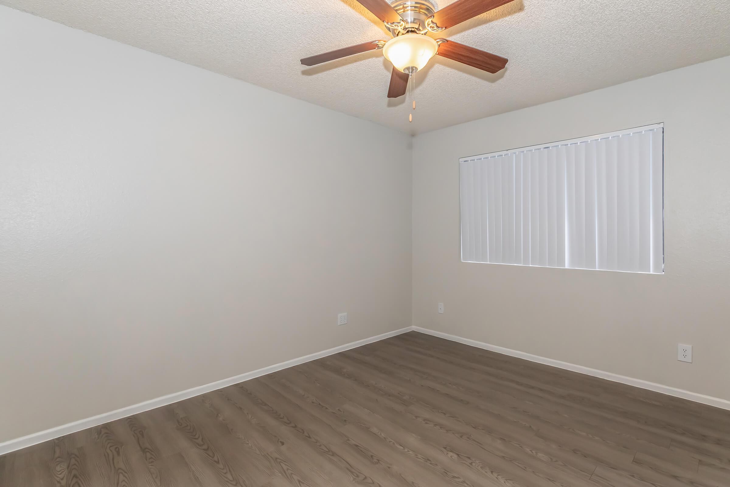 Empty room featuring light-colored walls, a ceiling fan with wooden blades, and a window with vertical blinds. The floor has a wood-like appearance, adding warmth to the space. Ideal for various uses, the room offers a blank canvas for personalization.
