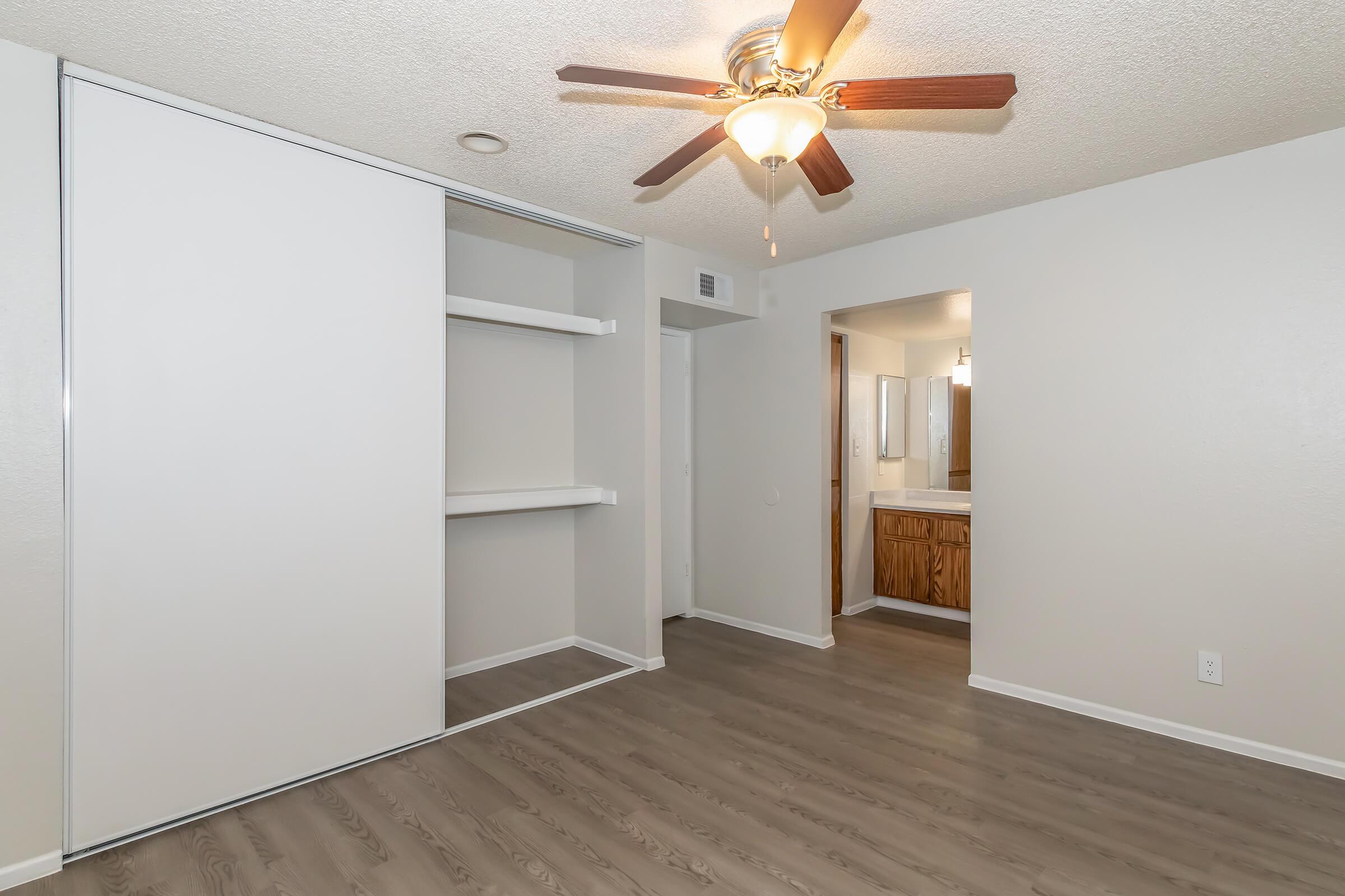 A well-lit bedroom featuring light-colored walls and a ceiling fan. There's a sliding door closet on one side and an open doorway leading to a bathroom on the right. The floor has a modern wood-like finish, creating a cozy and inviting atmosphere.
