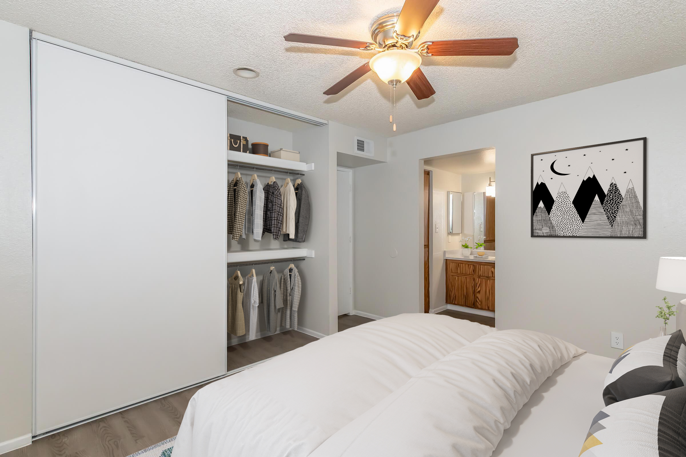 Modern bedroom featuring a king-size bed with white bedding, a ceiling fan, a closet with organized clothing, and an adjoining bathroom. The walls are painted in neutral tones, and there's a mounted artwork depicting mountains. The floor is hardwood, adding warmth to the space.