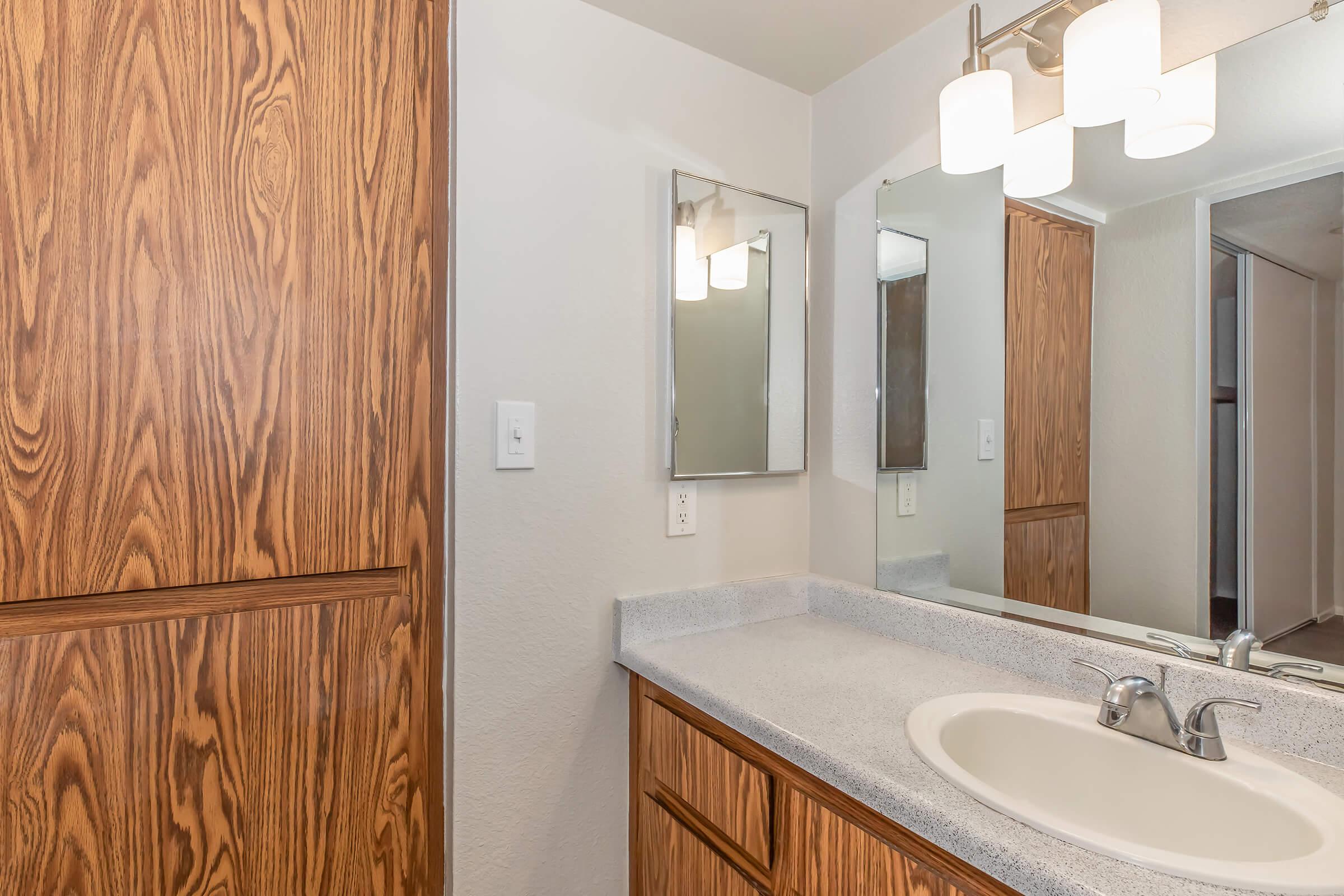 A modern bathroom featuring a light-colored countertop with a sink, wooden cabinetry, and a large mirror. The wall is painted in a neutral tone, and a light fixture with three bulbs is mounted above the mirror. A wooden door is visible, adding a warm touch to the space.