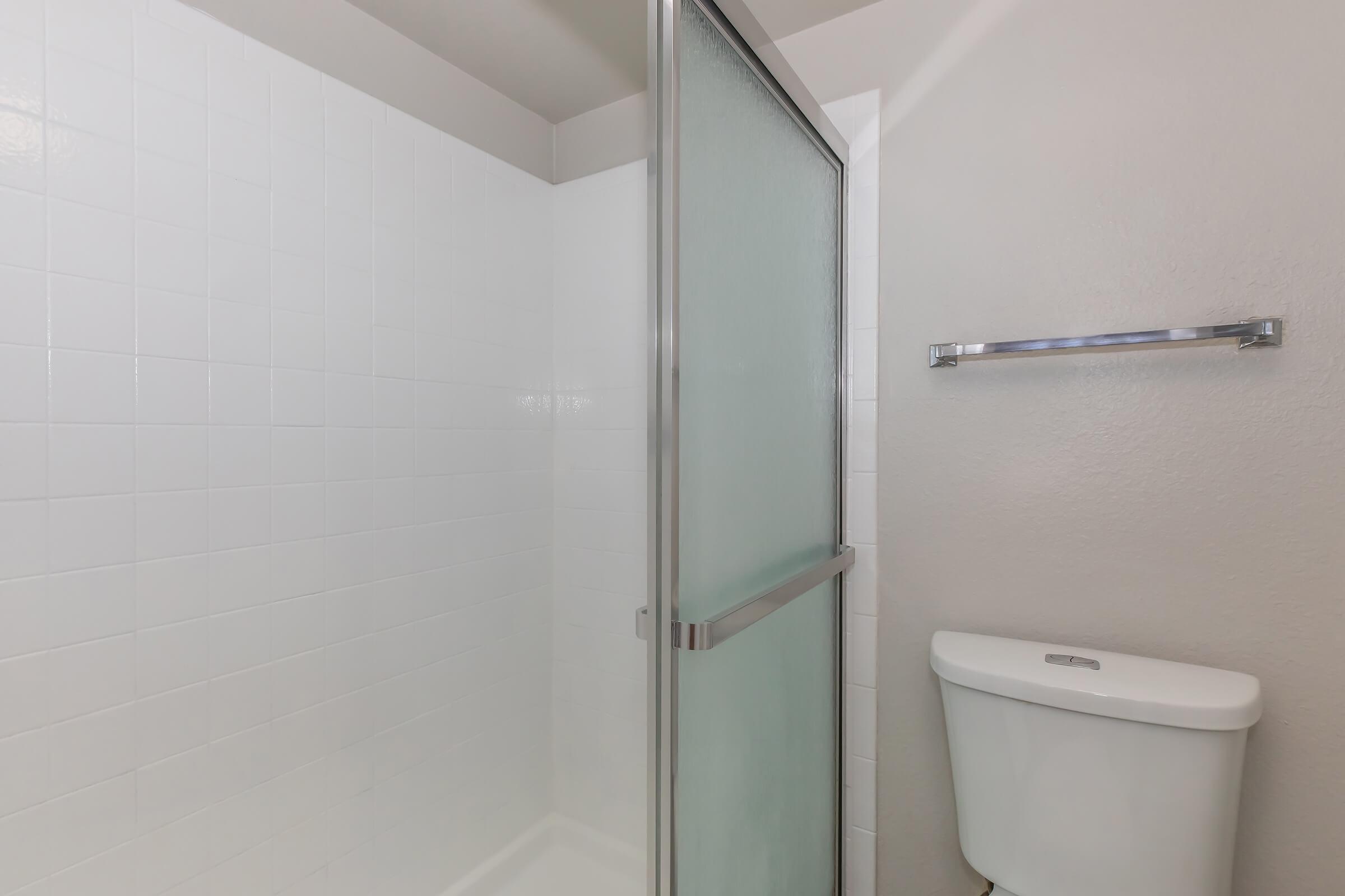 A clean, minimalist bathroom featuring a glass shower enclosure and a white toilet. The walls are tiled in white, and there is a silver towel bar mounted on the wall. The space is bright and well-lit, creating a fresh and modern aesthetic.