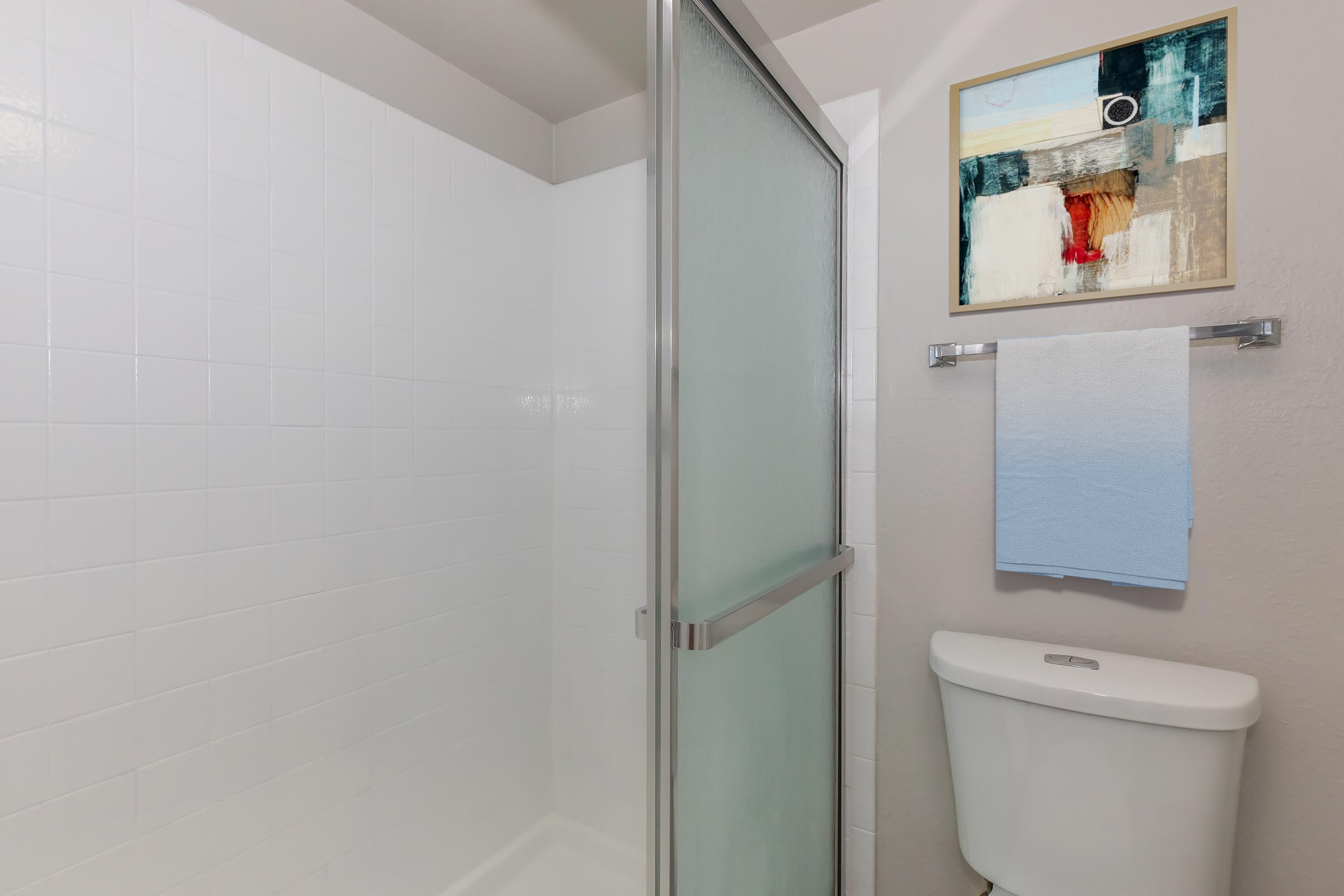 A clean and modern bathroom featuring a glass shower enclosure, a white toilet, and a wall-mounted towel rack with a blue towel. Above the towel rack, there is a framed piece of abstract art. The walls are light-colored and the space appears bright and minimalist.