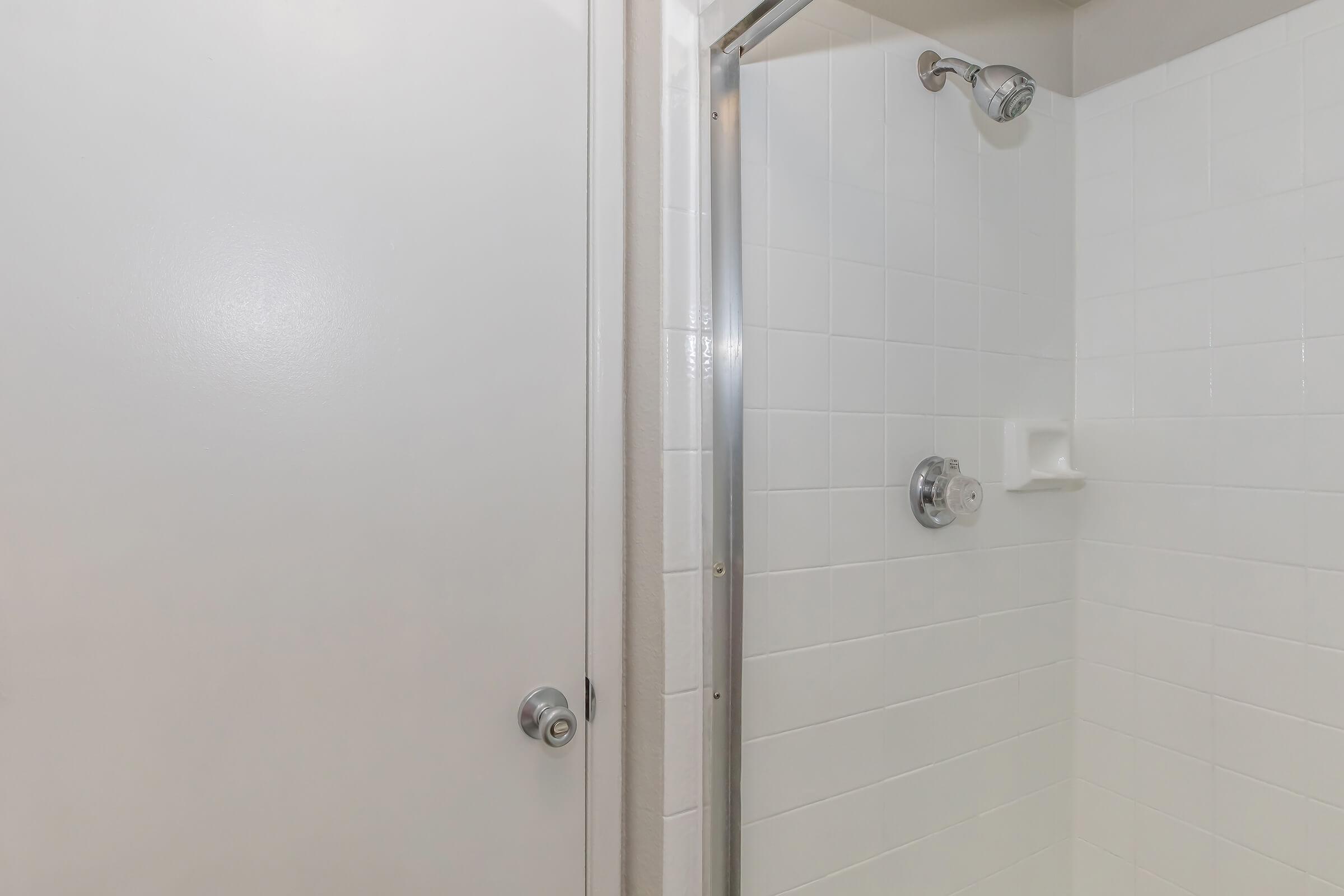 A clean and modern shower space featuring a glass door with a silver handle, white tiled walls, a showerhead mounted on the wall, and a small shelf for toiletries. The shower area is well-lit and appears freshly maintained, with a plain white door adjacent to the shower entrance.