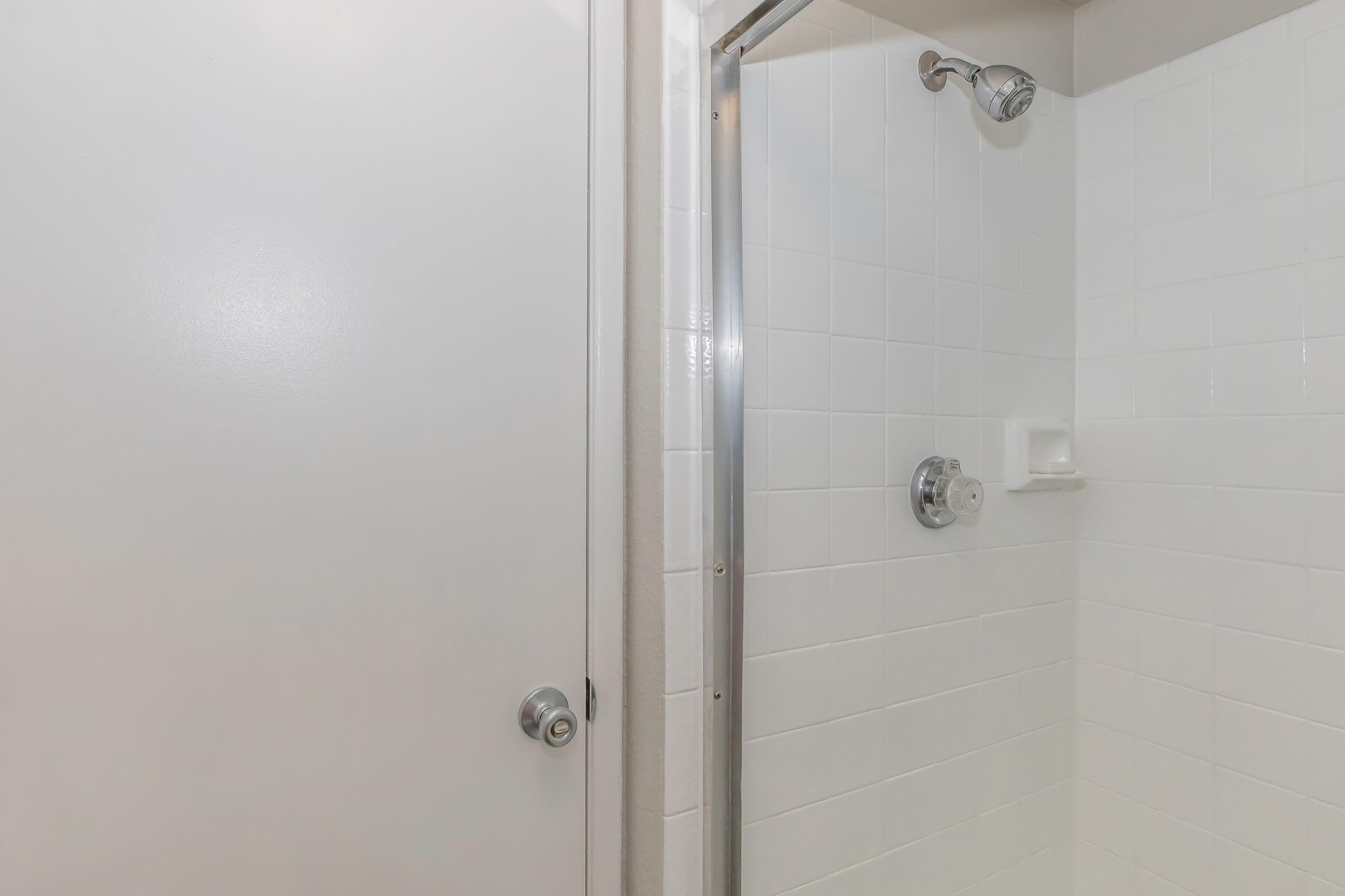 A clean, modern bathroom shower space featuring white tiled walls, a glass door, and a chrome showerhead. The door to the bathroom is partially visible, highlighting a simple design with a round doorknob. The overall aesthetic is bright and minimalist.