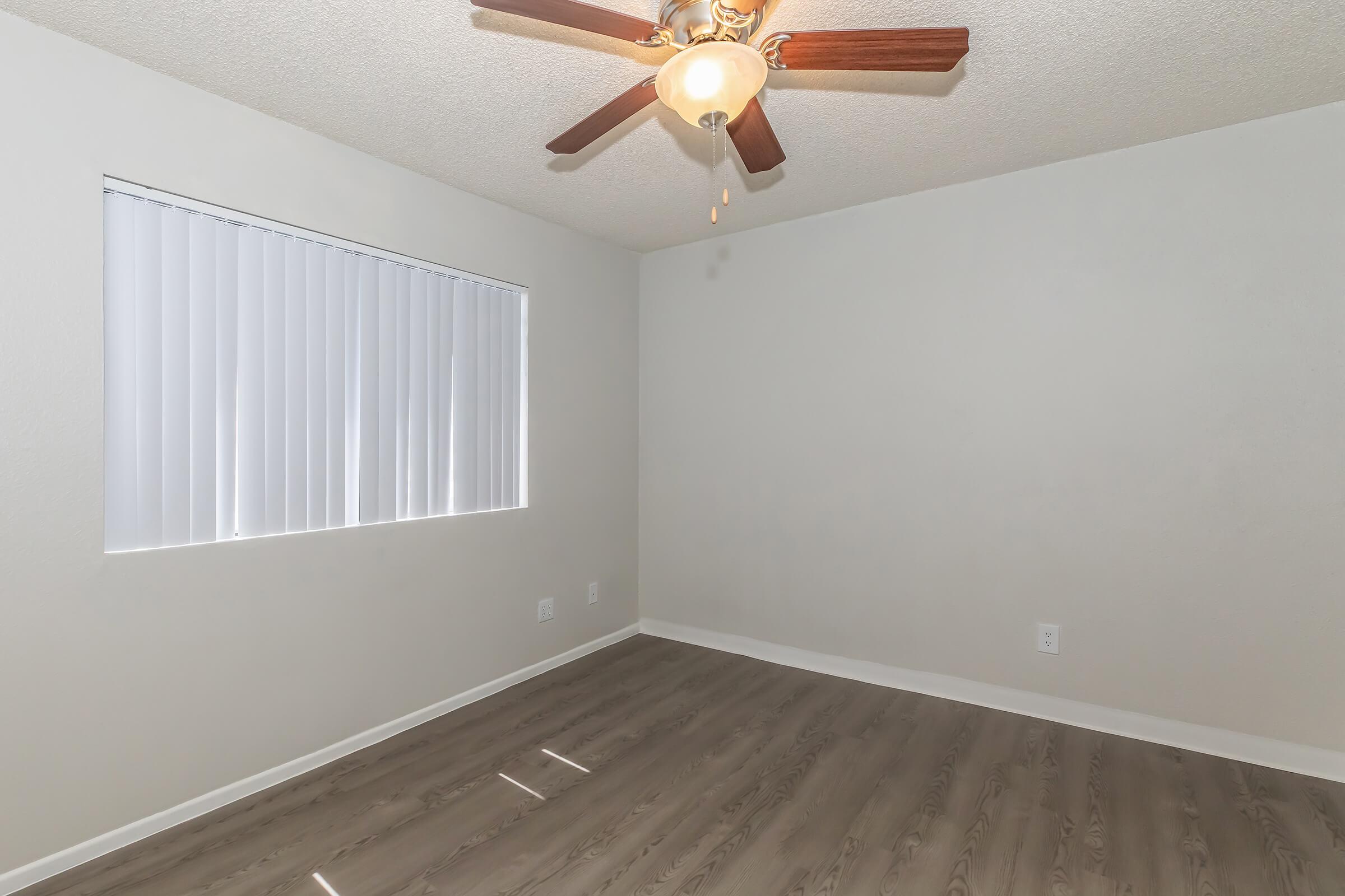 A well-lit, empty room featuring a ceiling fan with wooden blades. The walls are painted a light color, and there is a window with vertical blinds allowing natural light to enter. The floor is made of light-colored wood, giving the space a clean and modern look.