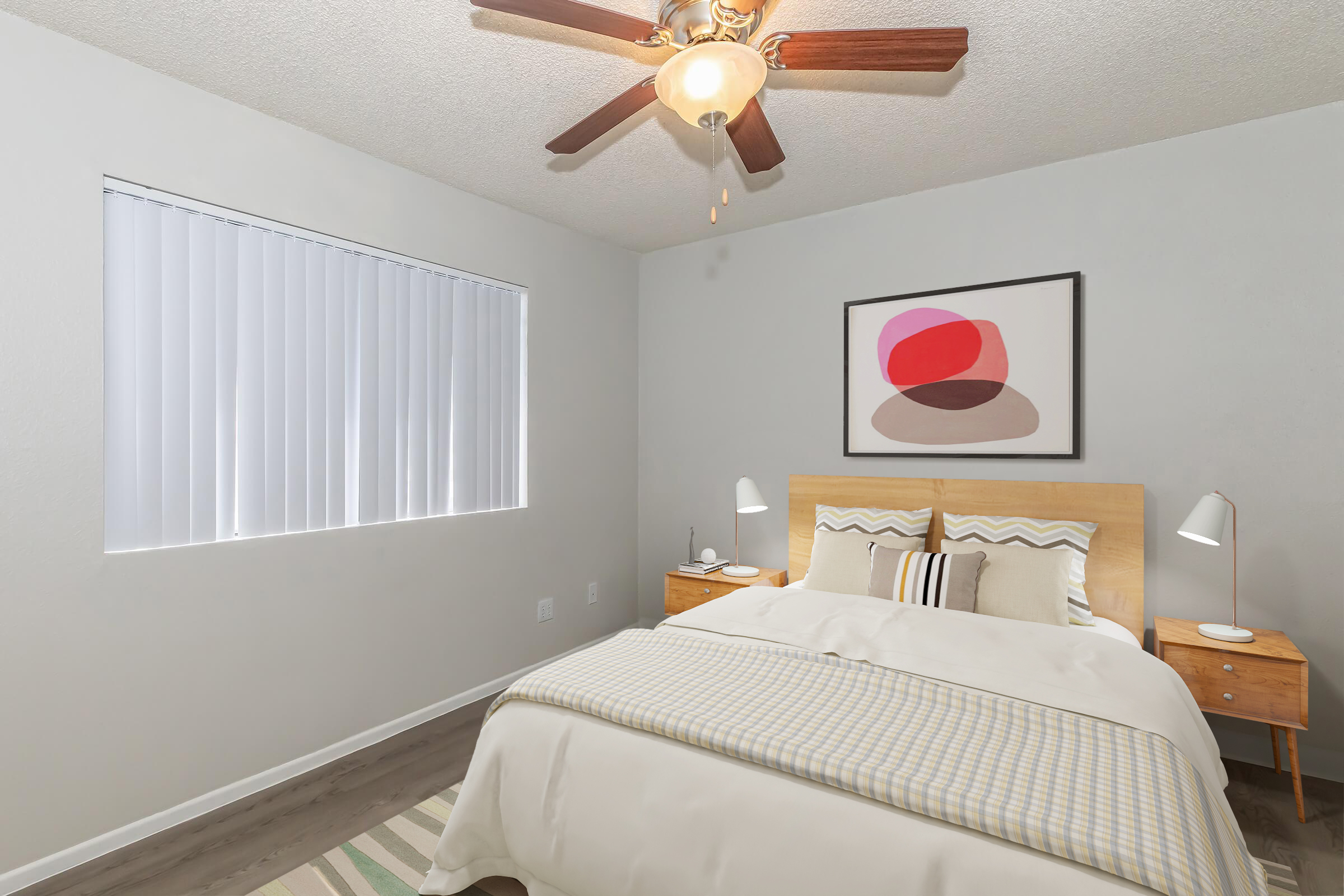 A cozy bedroom featuring a wooden bed with white bedding and decorative pillows. Two bedside tables with lamps are positioned on either side of the bed. A large window with white blinds allows natural light, and an abstract artwork hangs on the wall above the bed. The floor is covered with a subtle striped rug.