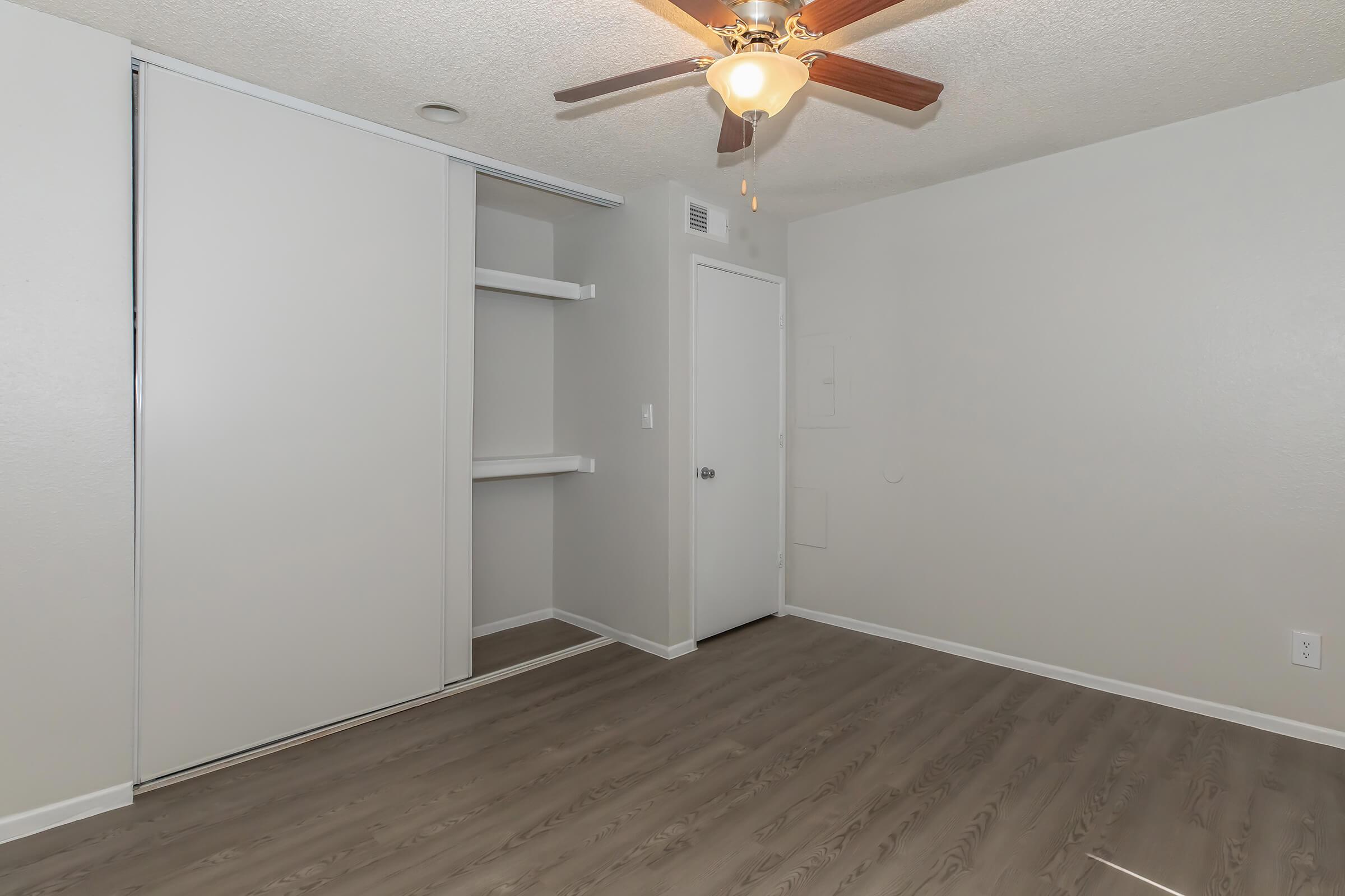 A clean, empty bedroom featuring a ceiling fan with light, light-colored walls, and laminate flooring. The closet has sliding doors, and there is a simple white door on one wall. The room is well-lit and has a spacious feel.