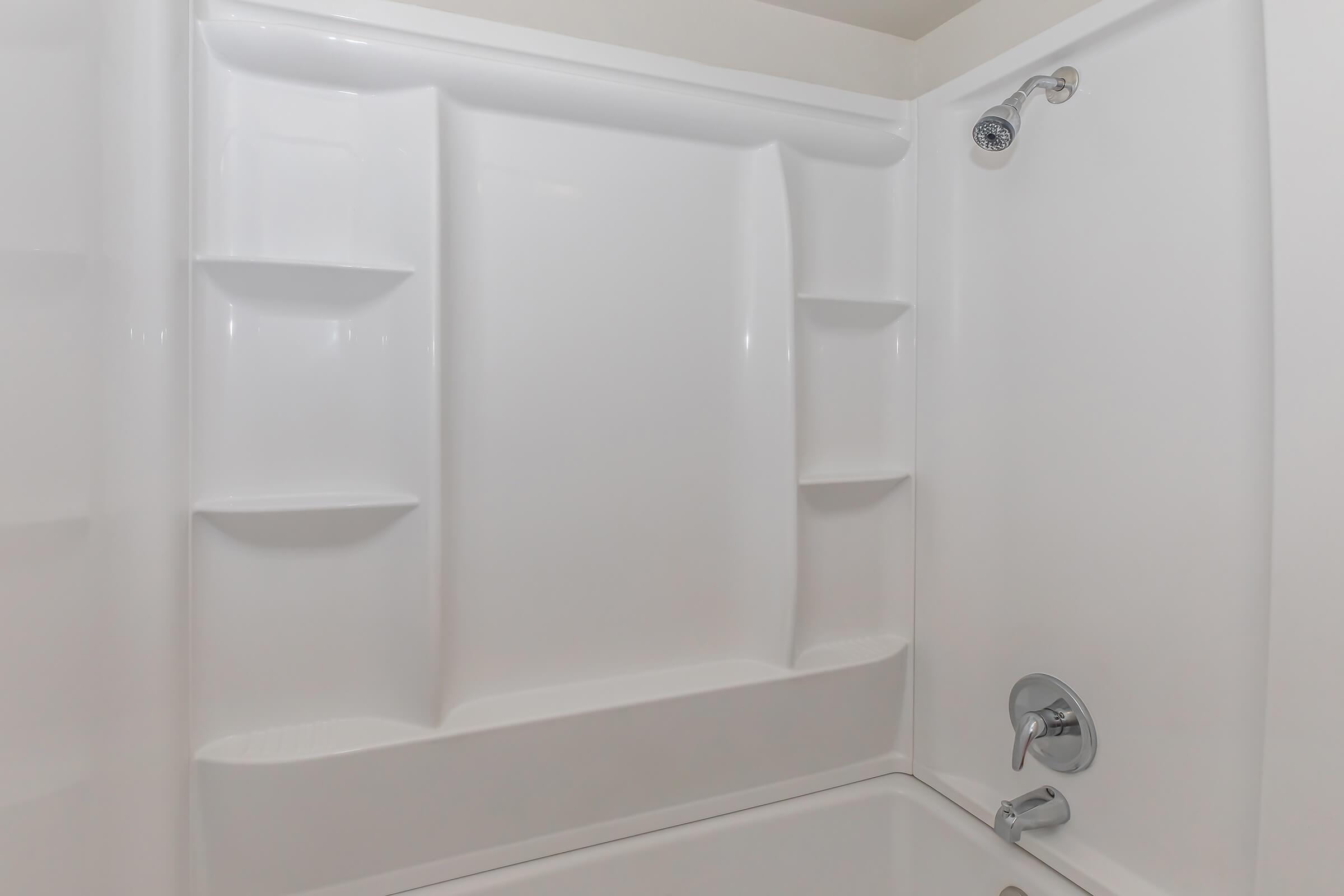 A clean, white shower stall featuring a molded plastic interior with built-in shelves for storing toiletries. The showerhead is installed above, and the faucet controls are visible on the wall. The overall design is sleek and minimalistic.