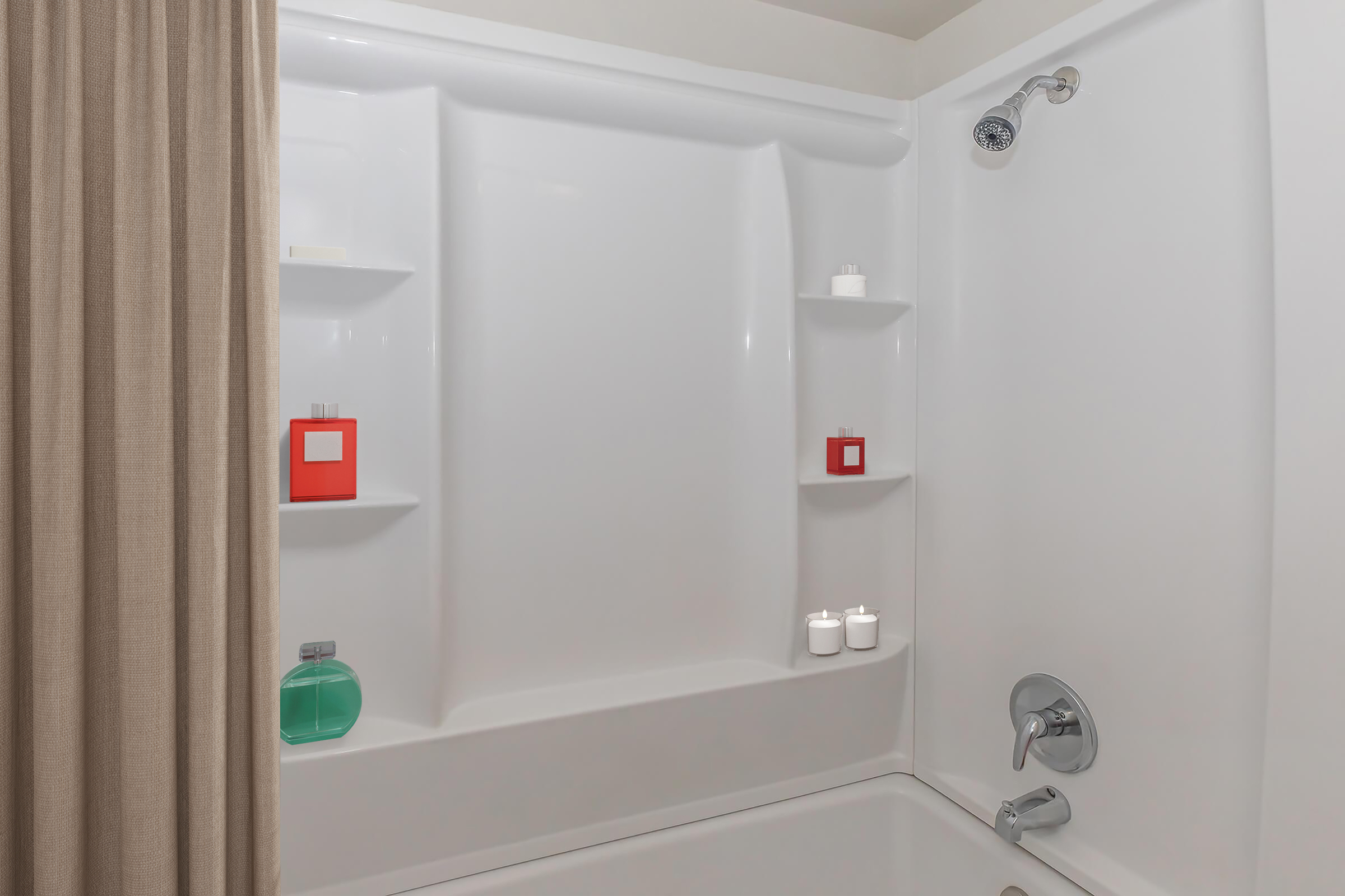 A clean, modern bathroom shower area featuring a white tub and walls. There are small shelves with decorative items, including red boxes and candles. A teal bottle is present on the tub's edge, and a water fixture is visible. A beige curtain partially separates the shower area.