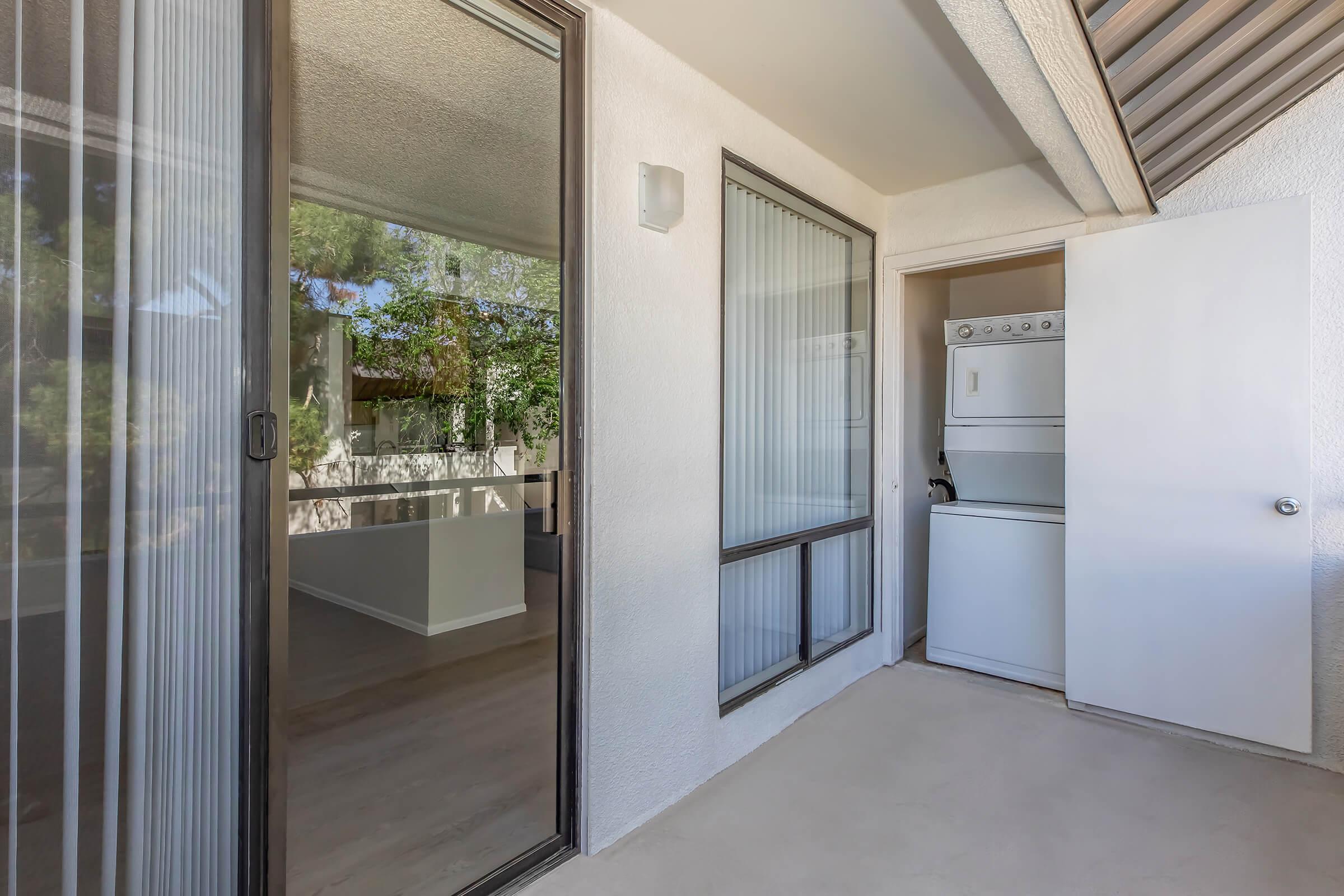 A view of a small balcony or patio area featuring a sliding glass door leading to an interior room. On one side, there is a stacked washer and dryer unit. The space is bright with natural light, and a green outdoor area is visible through the glass. The walls are light-colored, and the flooring appears to be a light laminate.