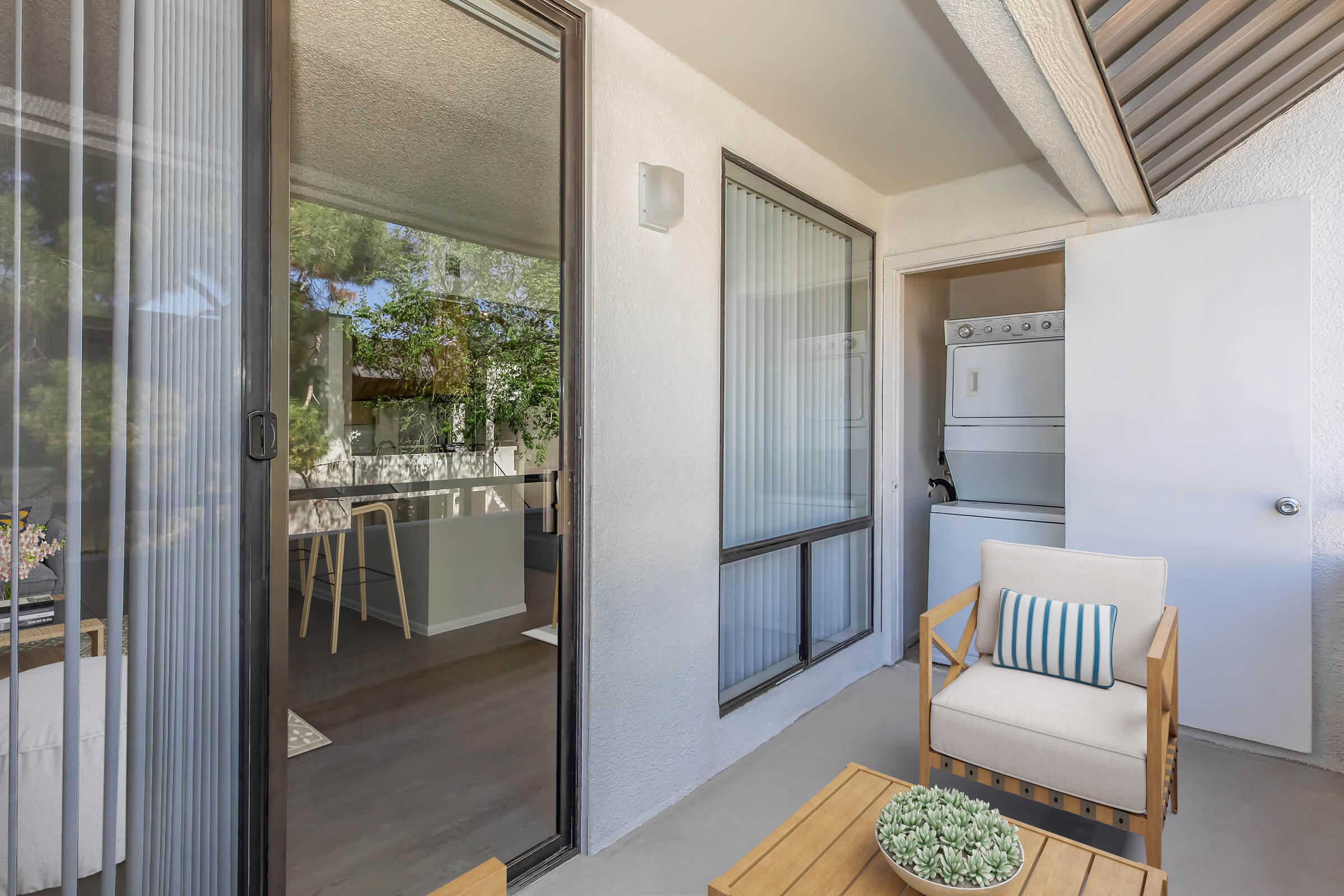 A small outdoor balcony featuring a beige armchair with a striped cushion and a wooden table. In the background, a glass door leads to a bright room and a laundry area with a washer and dryer. The space has a peaceful atmosphere with natural light and greenery visible through the windows.
