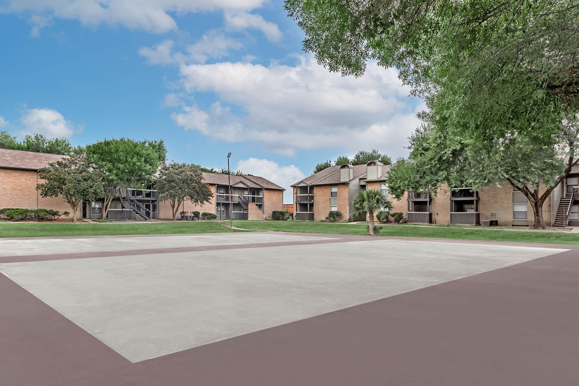 View of a residential area featuring several two-story apartment buildings surrounded by grassy lawns and trees. A concrete area in the foreground suggests recreational space, with cloudy blue skies overhead. The scene conveys a peaceful suburban environment.