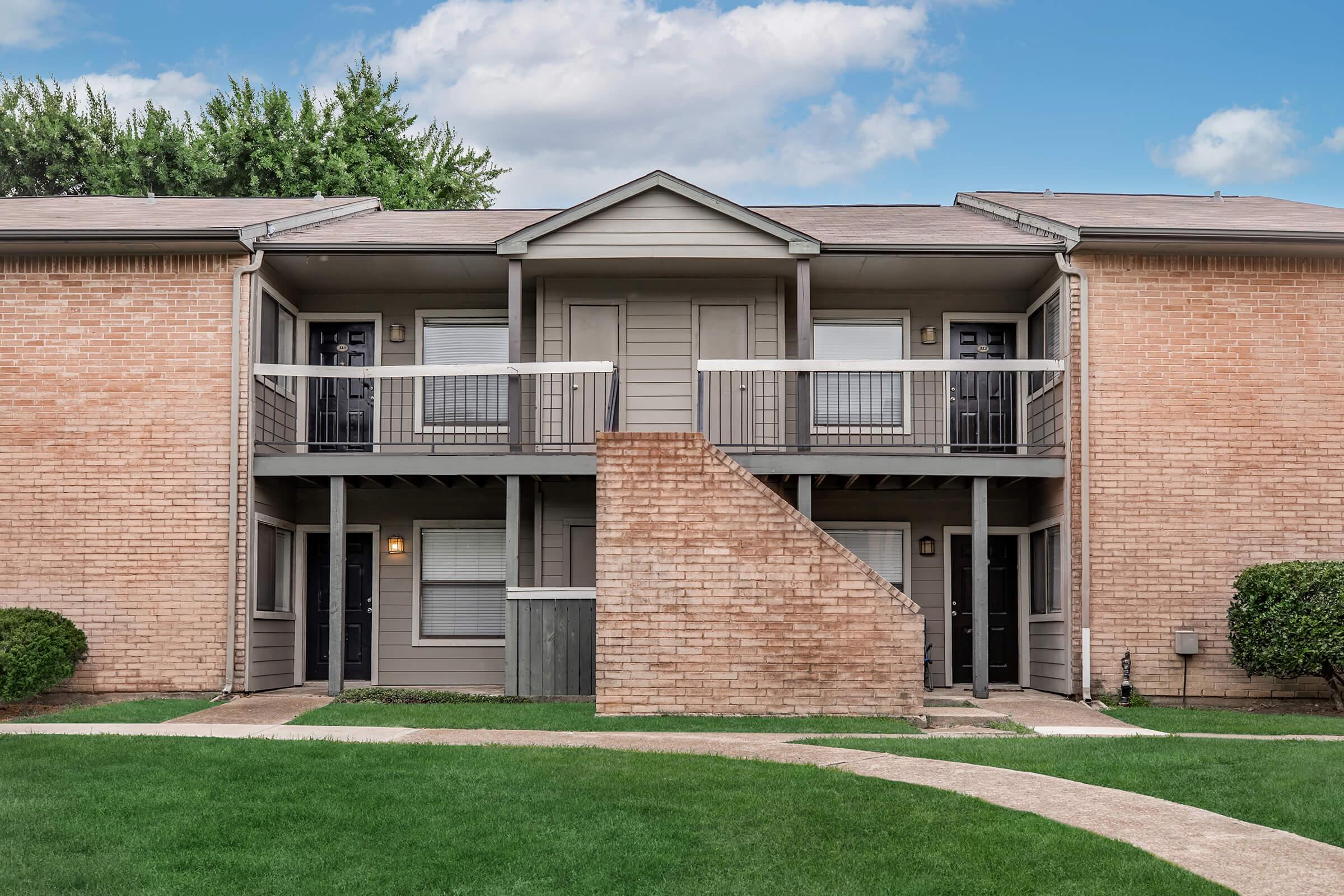 Two connected apartment units in a multi-family building, featuring a light brick exterior and two balconies with black railings. A small staircase leads from the ground level up to the balconies. The area is surrounded by manicured green grass and there are cloud-dotted skies above.