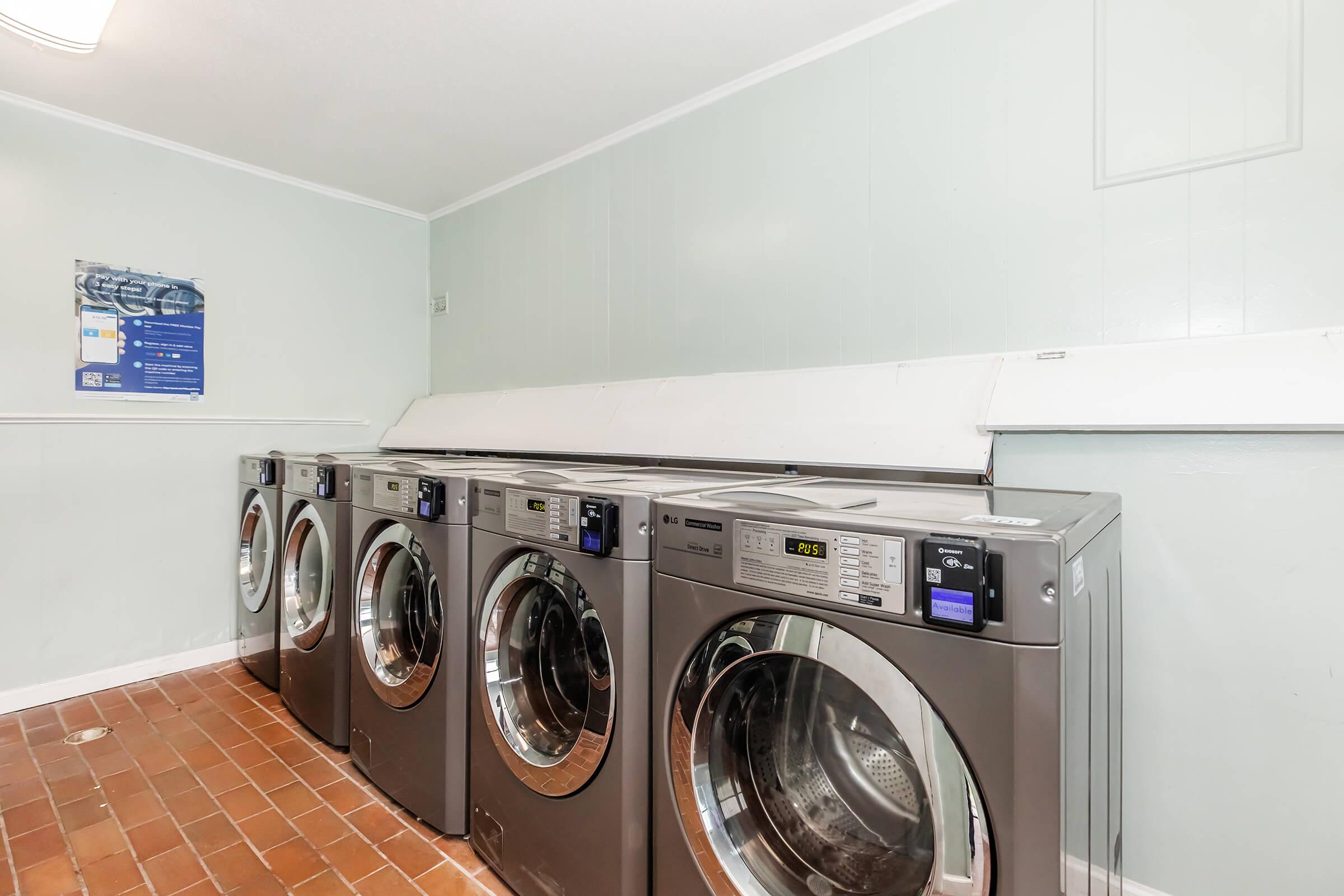 Four industrial washing machines lined up against a light green wall in a laundry room. The machines have clear glass doors and digital displays. A poster on the wall provides laundry instructions. The floor is made of brown tiles, contributing to a clean and functional atmosphere.