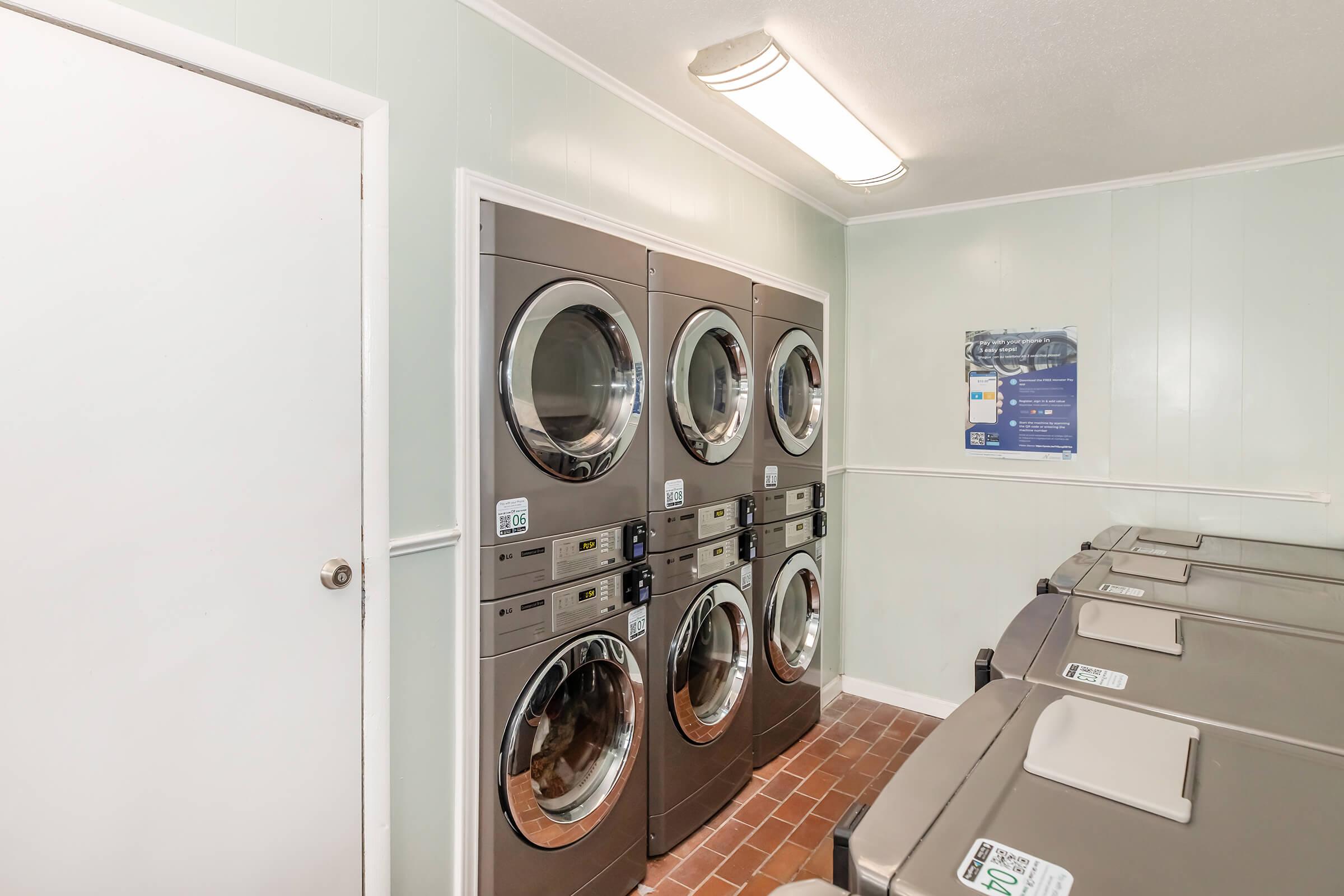 A laundry room featuring stacked silver dryers and large commercial washing machines along a light green wall. A white door is visible on the left side, and a poster on the wall offers laundry instructions or promotions. The floor is tiled in a reddish-brown color.
