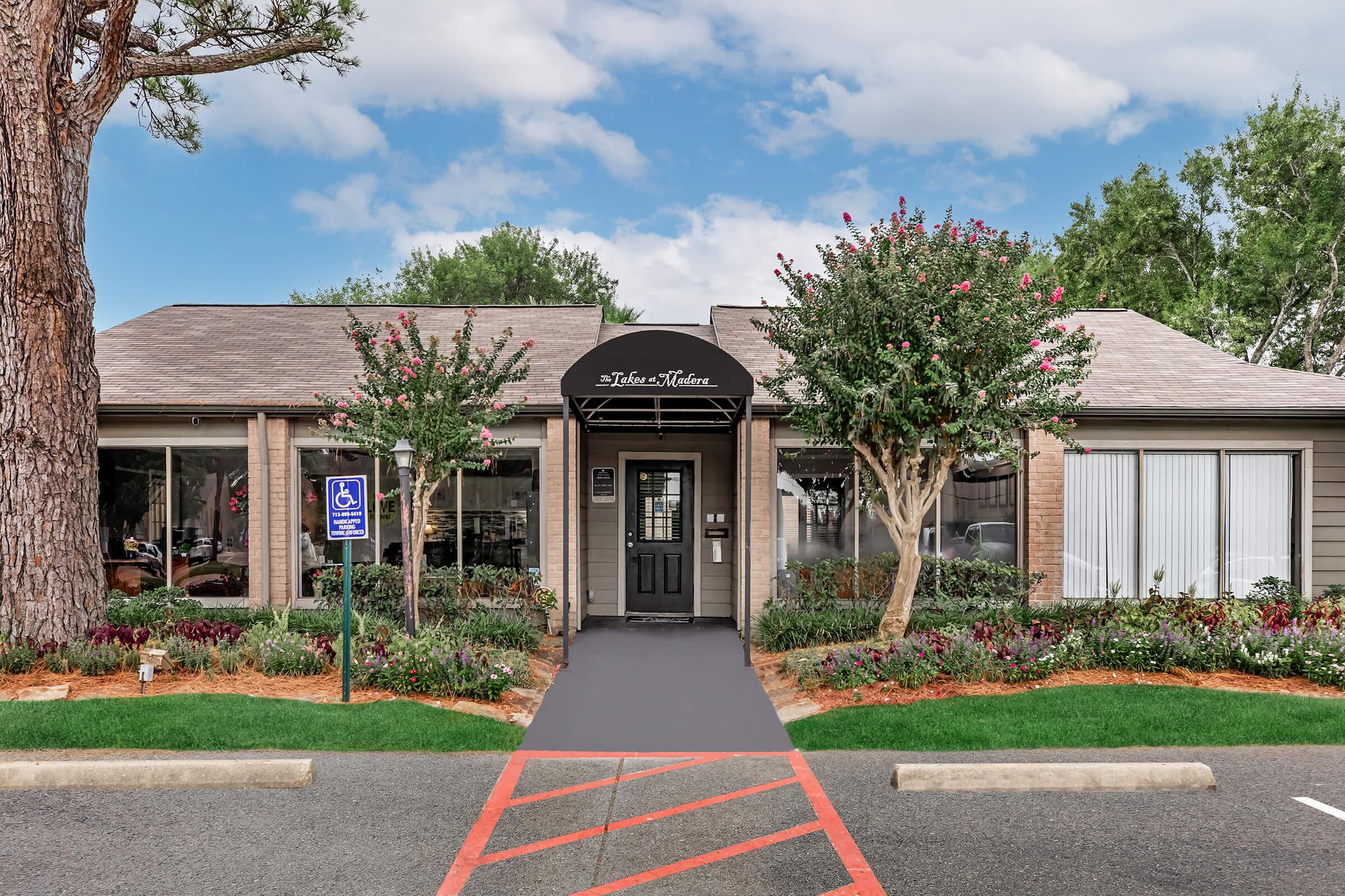 Exterior of a single-story building with a sign that reads "Theater Kitchen." The facade features large windows, a black door, and decorative landscaping including trees and flowering plants. A paved walkway leads to the entrance, with accessible parking visible to the left. The sky is partly cloudy.