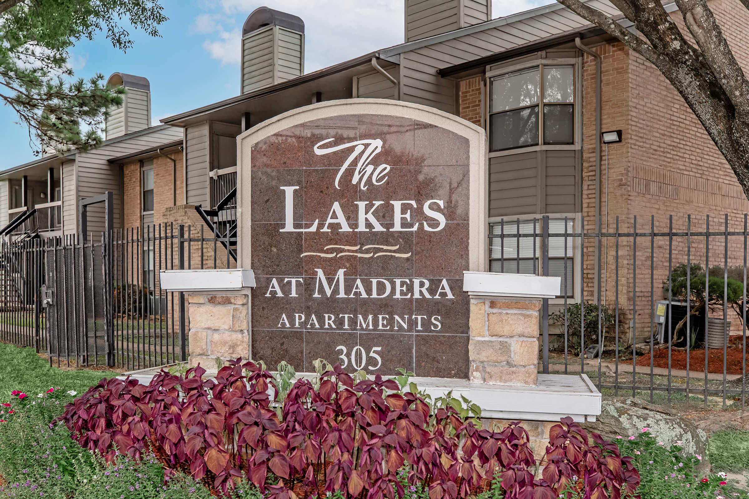 Sign for "The Lakes at Madera Apartments" with the address 305, surrounded by greenery and flowers in front of a multi-story apartment building. The sign features a decorative design and is positioned near a fence.
