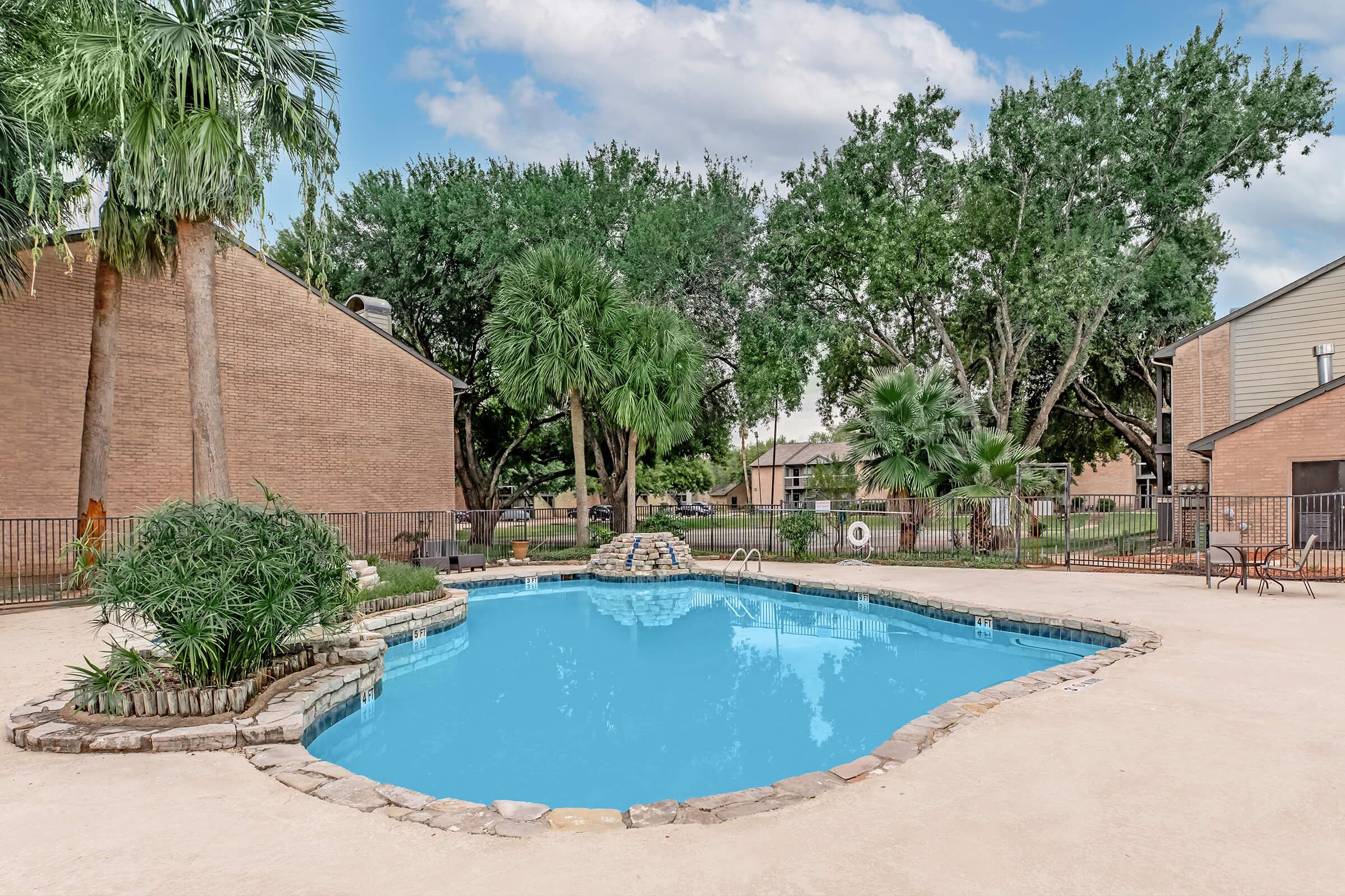 A serene swimming pool surrounded by lush greenery and palm trees. The pool features a rock border and a small stone waterfall, with lounge chairs nearby. In the background, there are two residential buildings and a clear blue sky, creating a peaceful outdoor retreat.
