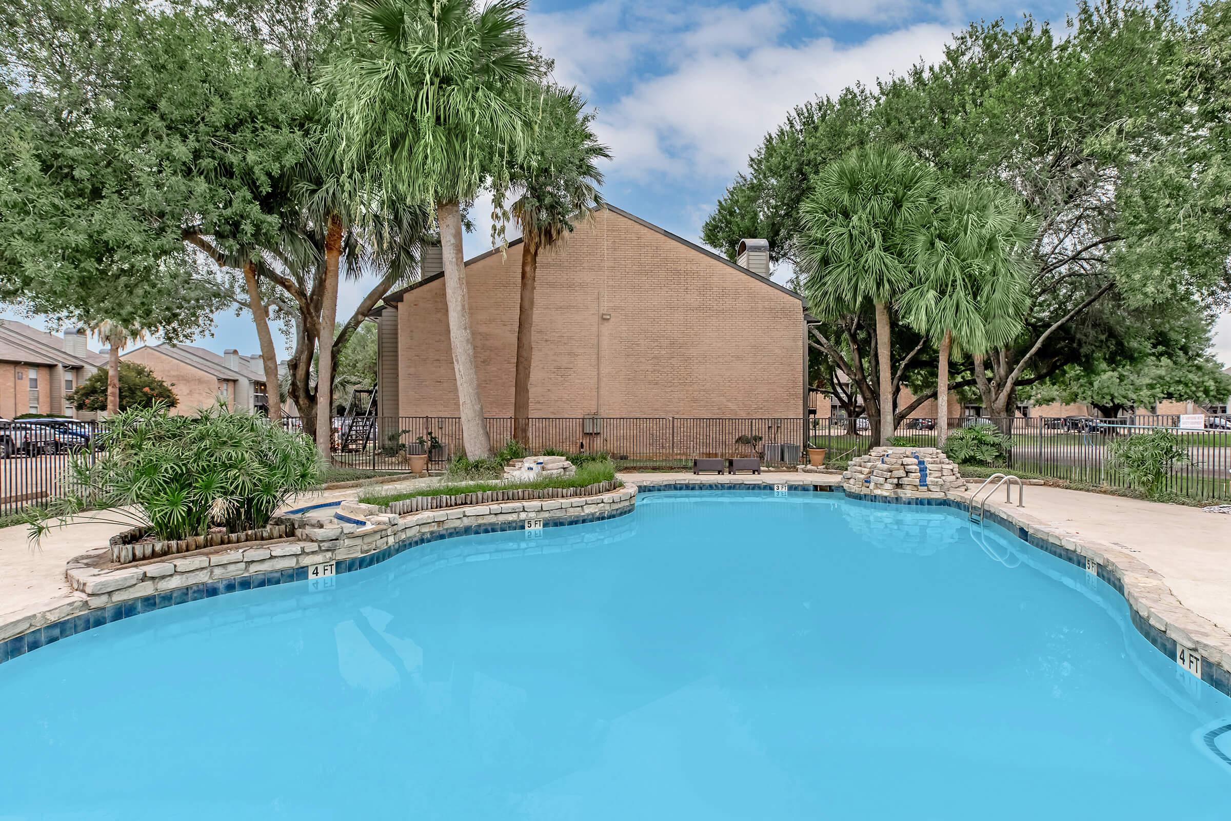 A clear blue swimming pool surrounded by palm trees and landscaped greenery. In the background, there is a beige building partially visible. The pool area features a stone edge and a few lounge chairs. The sky is partly cloudy, creating a relaxed atmosphere.