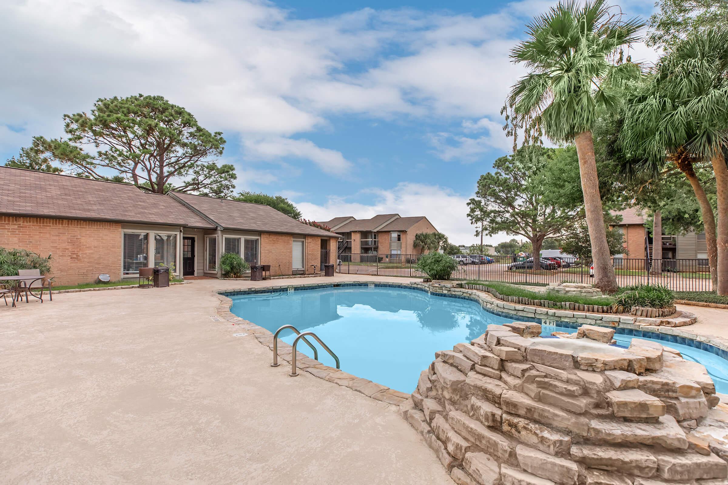 A serene outdoor pool area featuring a curved pool surrounded by a stone deck. Palm trees and greenery provide a relaxing atmosphere. Nearby, a collection of brown brick buildings adds to the charm, with clear blue skies overhead enhancing the tranquil setting.
