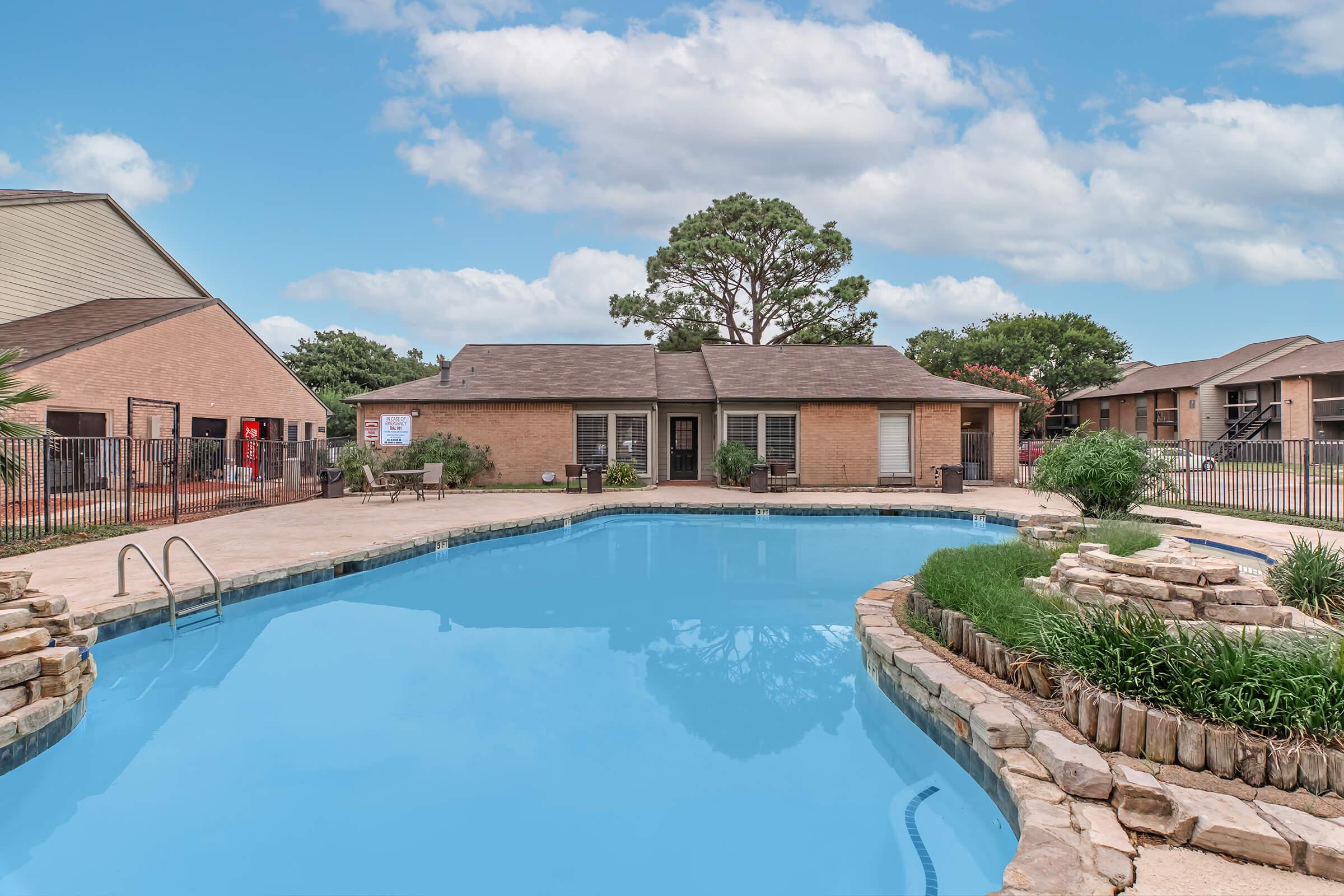A swimming pool surrounded by a stone patio, featuring a small waterfall and lush greenery. In the background, there are residential buildings and a clear blue sky with clouds. The setting is inviting and well-maintained, ideal for relaxation and leisure.