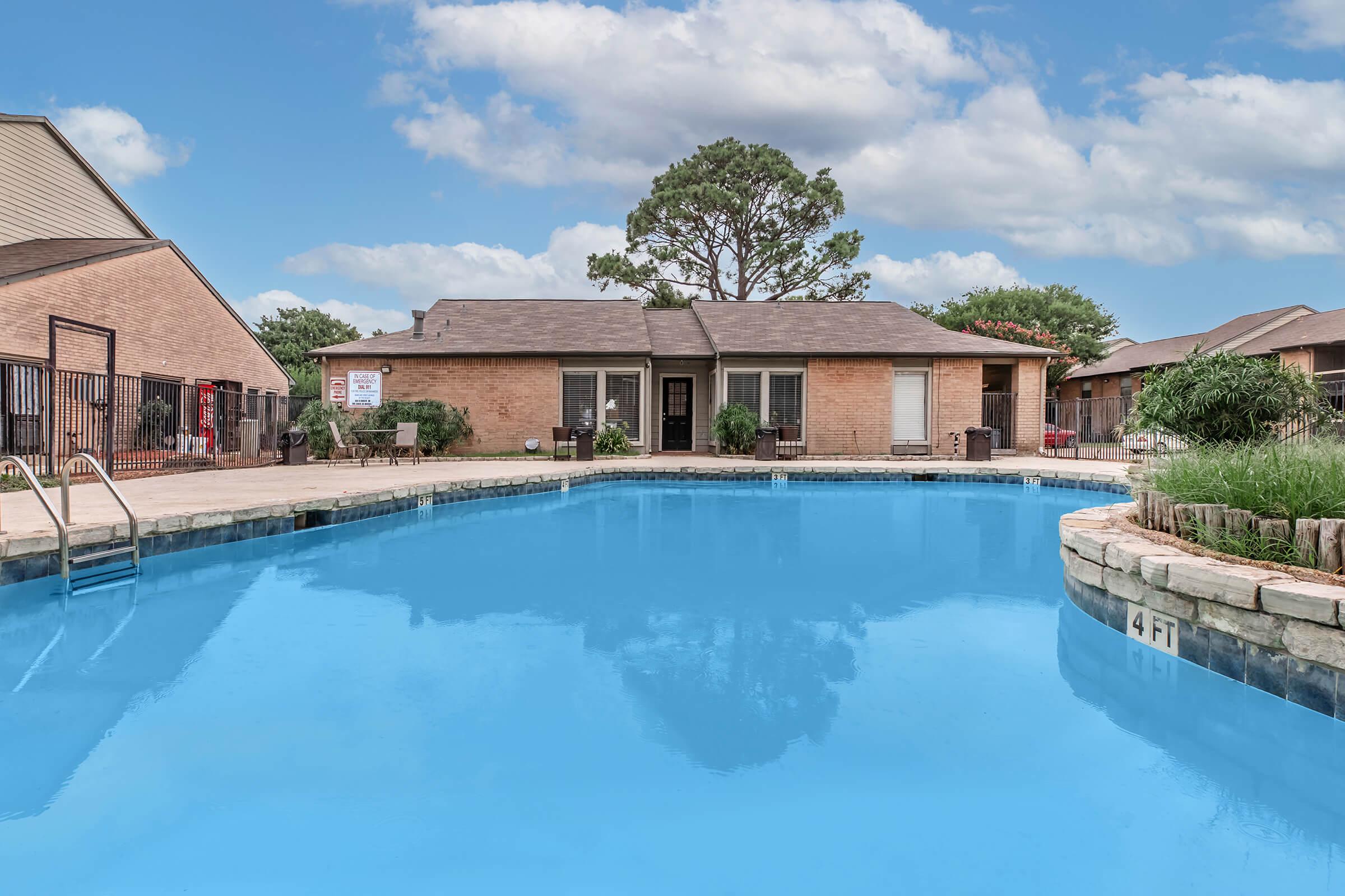 A large outdoor swimming pool with crystal-clear blue water, surrounded by a stone edge. There is a residential building in the background, framed by trees and greenery. The sky is partly cloudy, adding a serene atmosphere to the scene. A pool ladder is visible on the left side.
