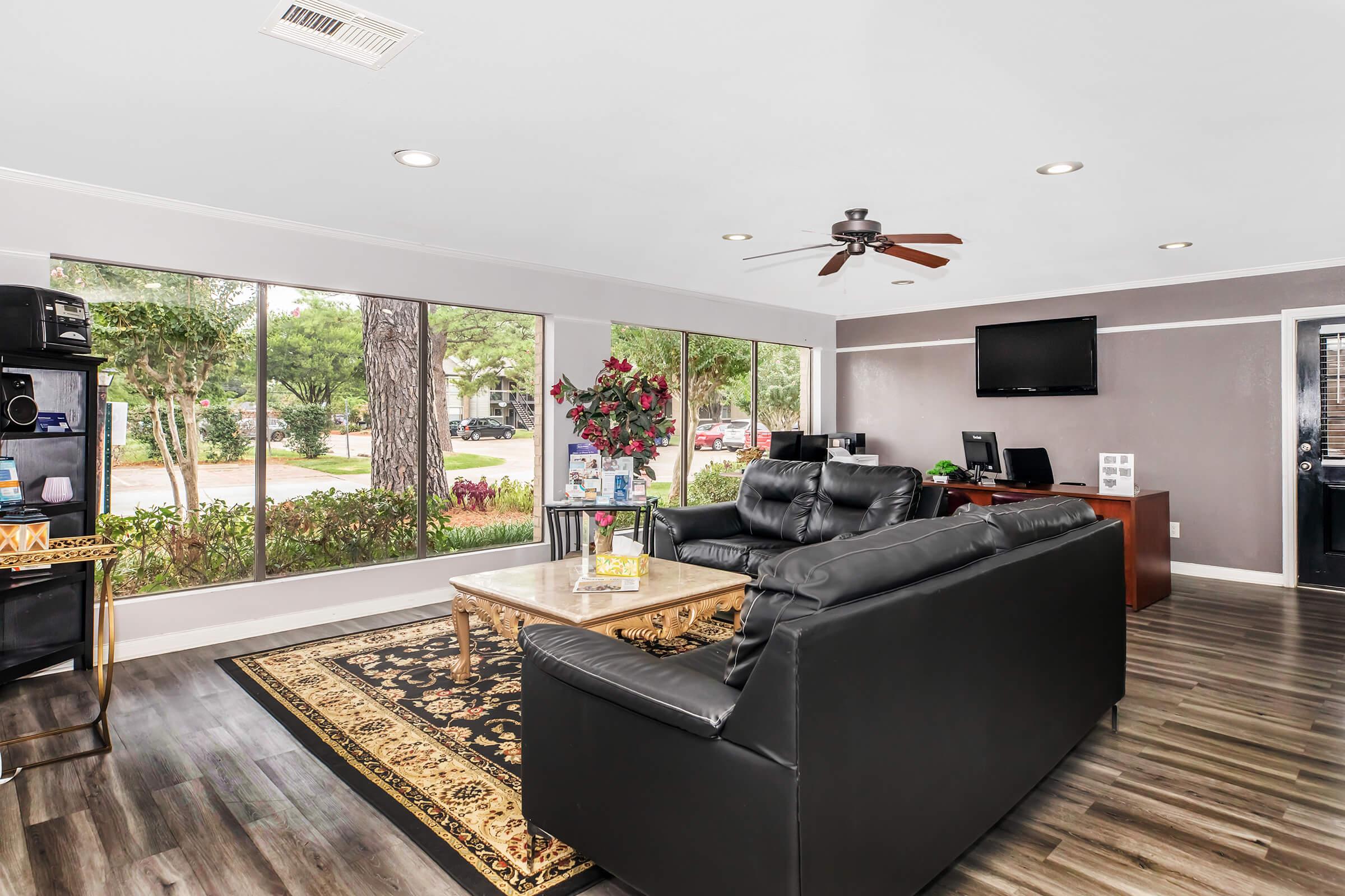 A modern lounge area featuring black leather sofas, a decorative coffee table, and a large window with views of greenery outside. The space includes a ceiling fan, a television mounted on the wall, and a reception desk with computer stations. Decorated with potted plants and a patterned rug.