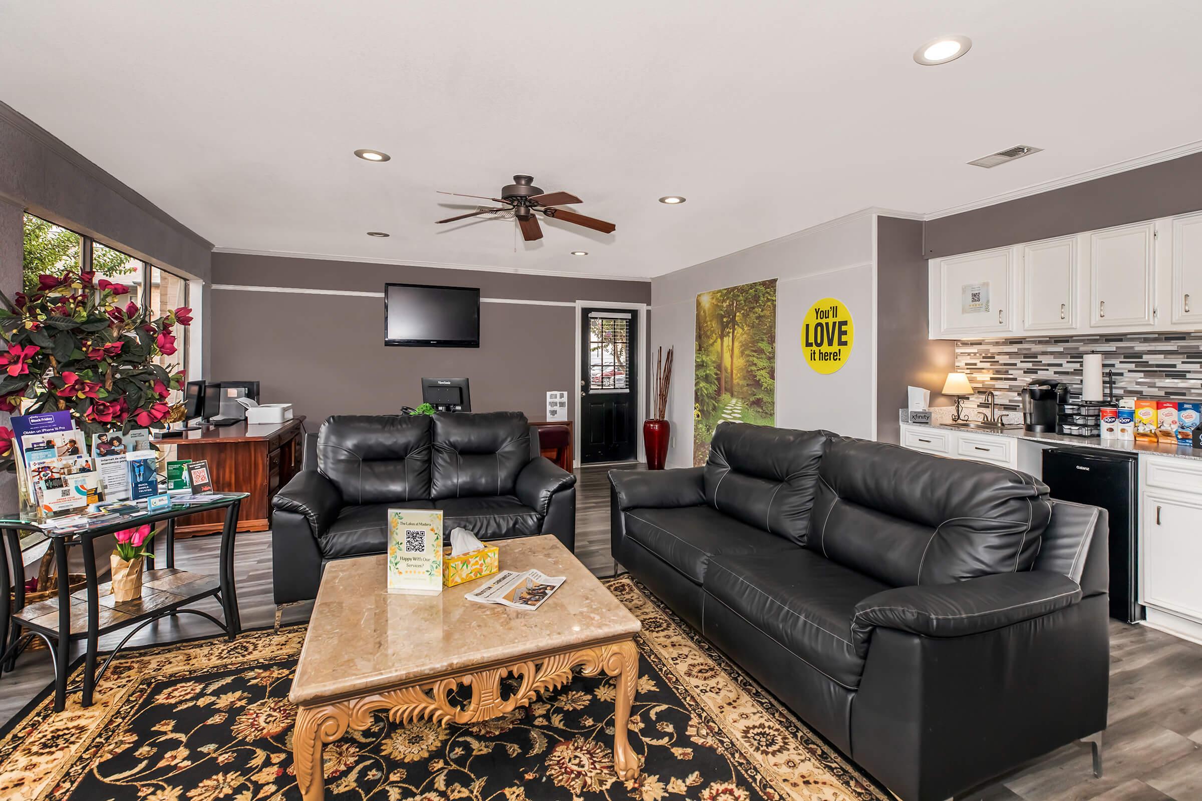 A welcoming lounge area featuring two black leather sofas, a coffee table, and a decorative rug. There's a reception desk in the background, a wall-mounted TV, and a kitchenette with cabinets. Bright floral plants and a wall mural add color to the space, creating a comfortable environment for guests.