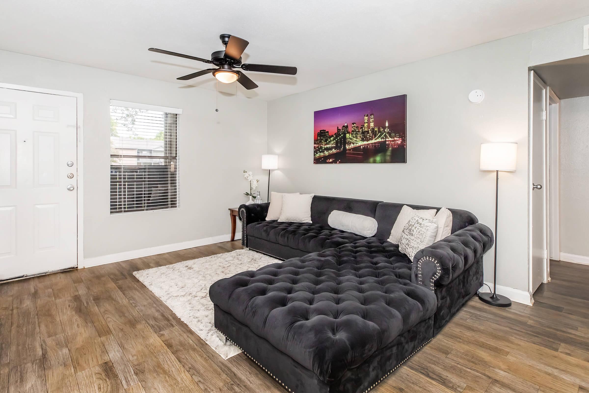 A cozy living room featuring a black tufted sectional sofa with decorative pillows, a large area rug, and wooden flooring. A ceiling fan hangs above, and there's a wall art piece depicting a city skyline at night. Natural light streams in through a window, and a small side table with a lamp adds to the ambiance.