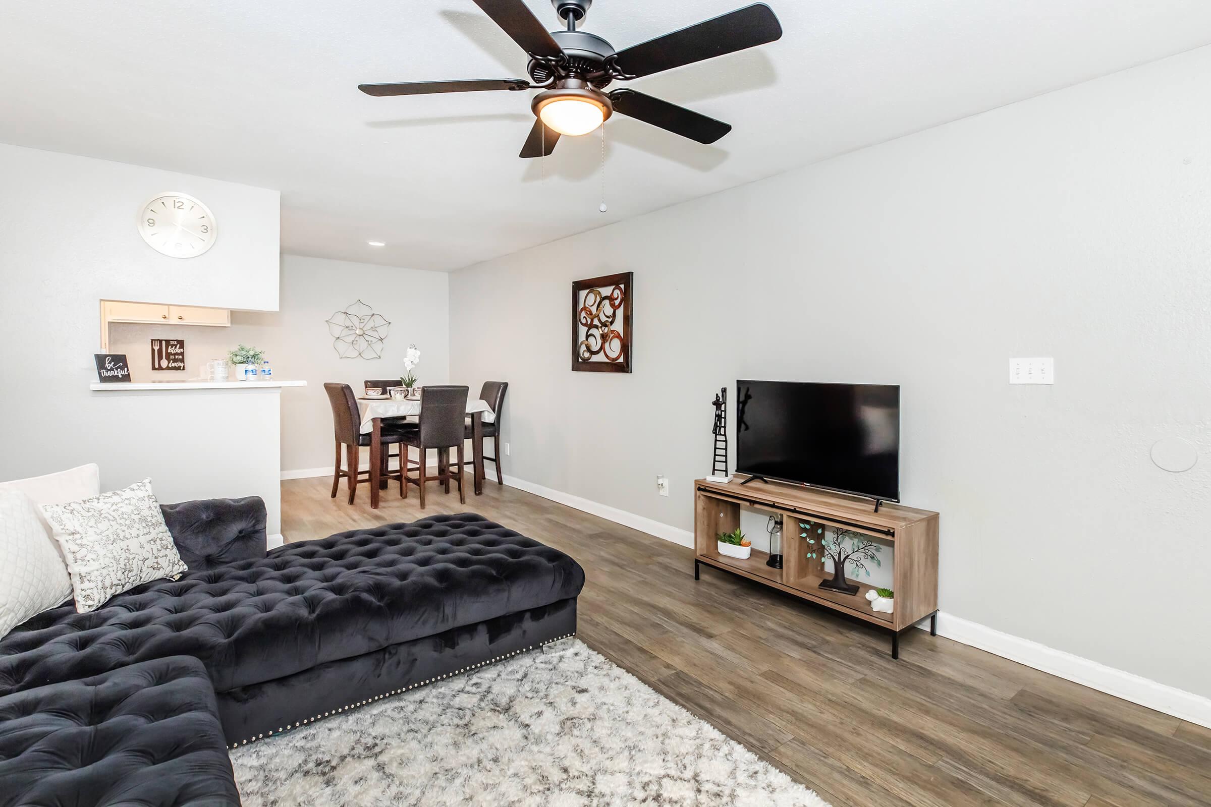 A modern living room featuring a dark velvet sectional sofa, a decorative area rug, a wall-mounted TV on a wooden stand, and a small dining area with a table and chairs. The walls are light gray, and there is a ceiling fan with light. Decorative elements include wall art and plants.