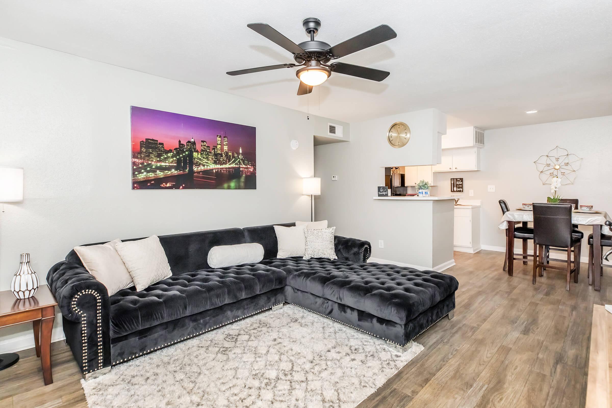A modern living room featuring a dark velvet sectional sofa with decorative pillows, a light-colored area rug, and contemporary lighting. A wall-mounted artwork depicts a city skyline at night. In the background, there's a dining area with a table and chairs, and a glimpse of a kitchen.
