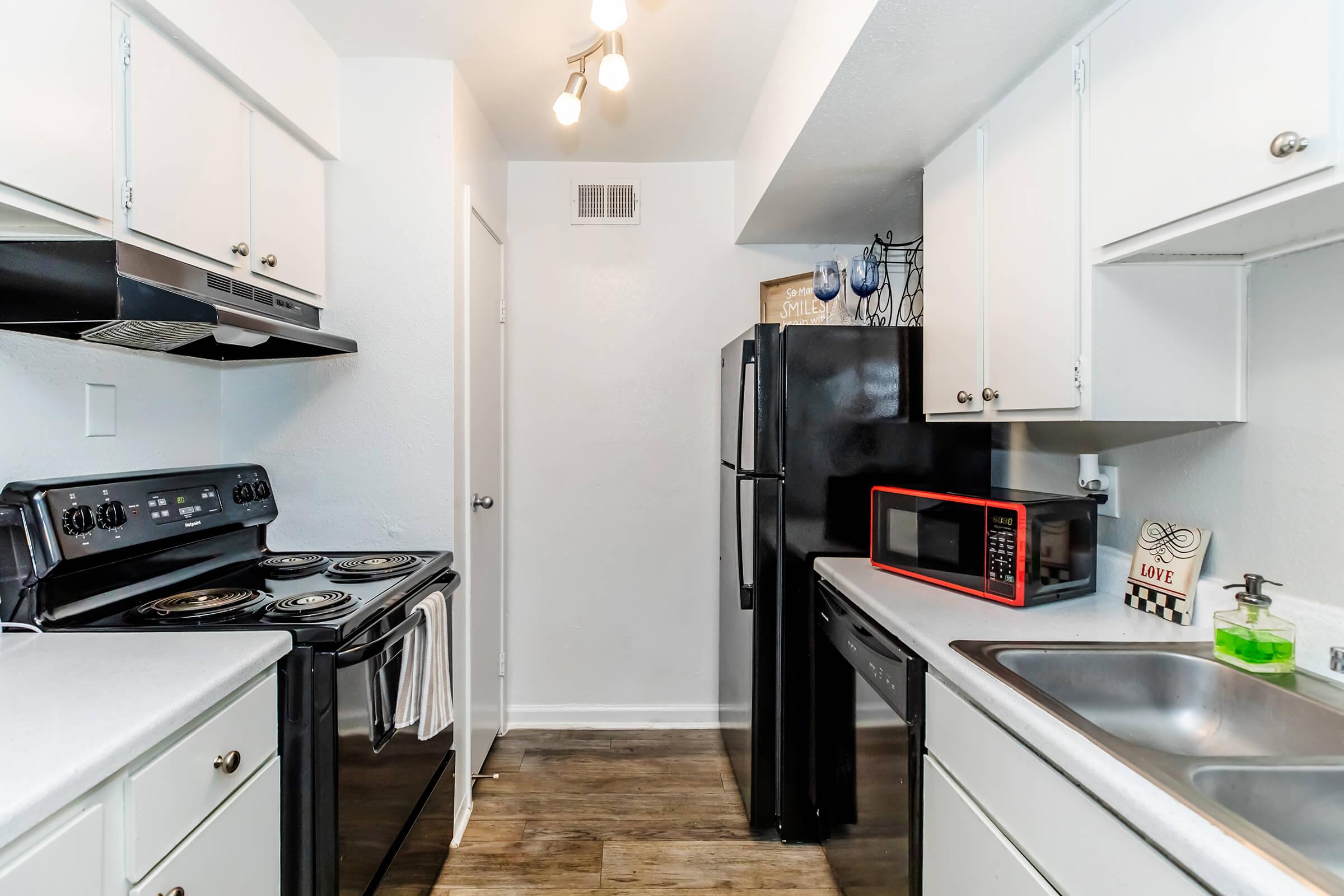 A modern kitchen featuring black appliances including a stove, refrigerator, and microwave. The cabinetry is white with a light countertop. There are overhead lights and a small decorative sign on the counter. The flooring is a wood-like laminate, giving the space a clean and contemporary look.