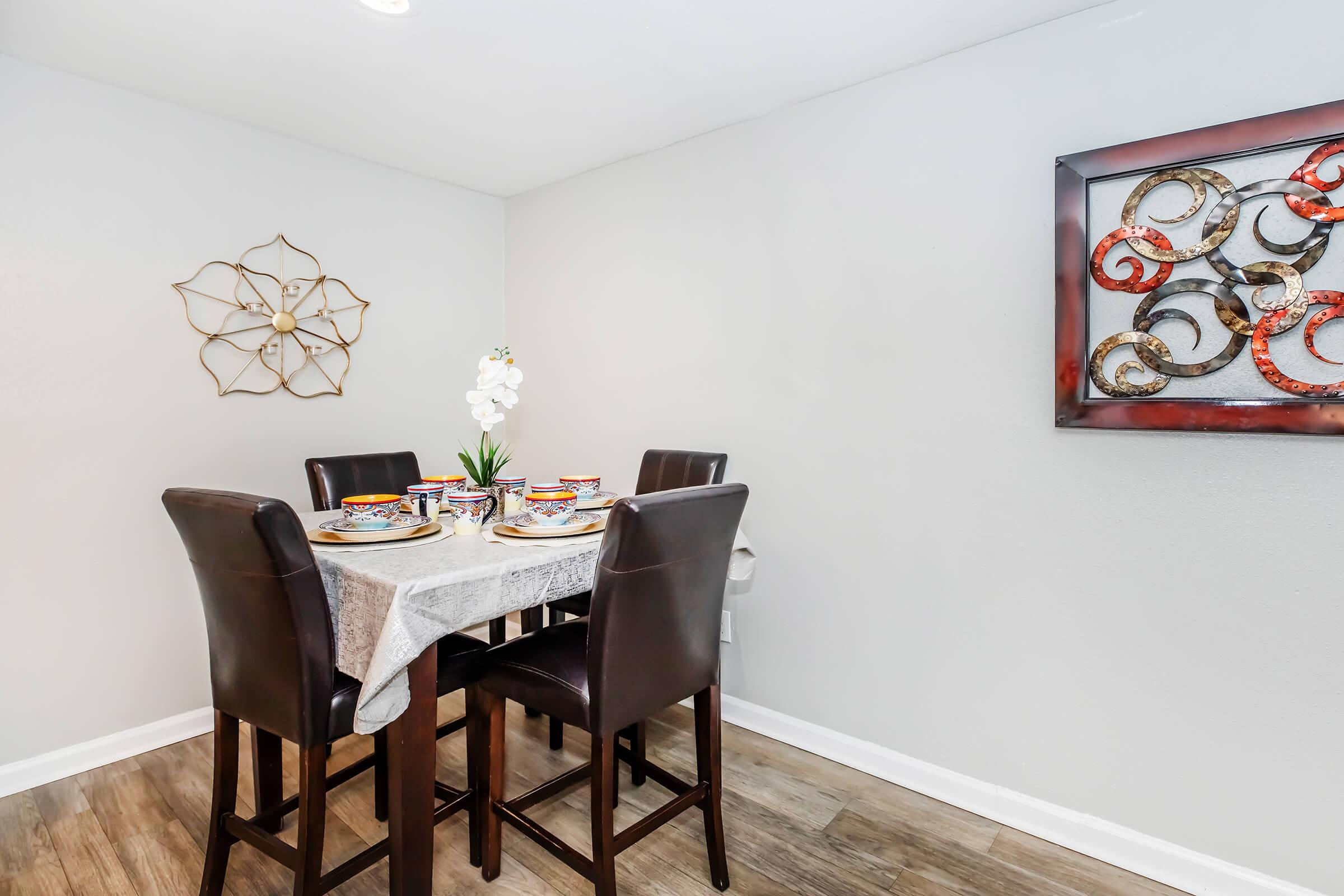 A stylish dining area featuring a round table set with colorful dishware and decorative plates, surrounded by four dark leather chairs. A gold wall decoration and a modern artwork piece enhance the ambiance, with a small plant and white flower centerpiece adding a touch of elegance.