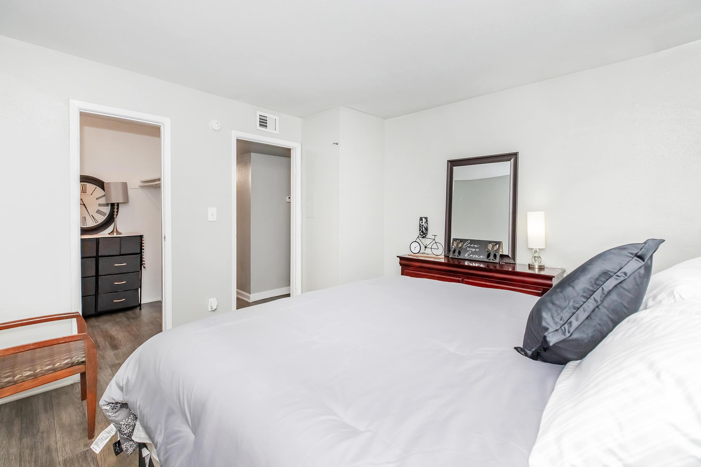 A clean and modern bedroom featuring a neatly made bed with gray bedding. There is a large mirror on a dresser, a wooden chair, and a stylish wall clock visible in the background. The room is well-lit with neutral wall colors and flooring, creating a cozy atmosphere. A closet door is partially open to the side.