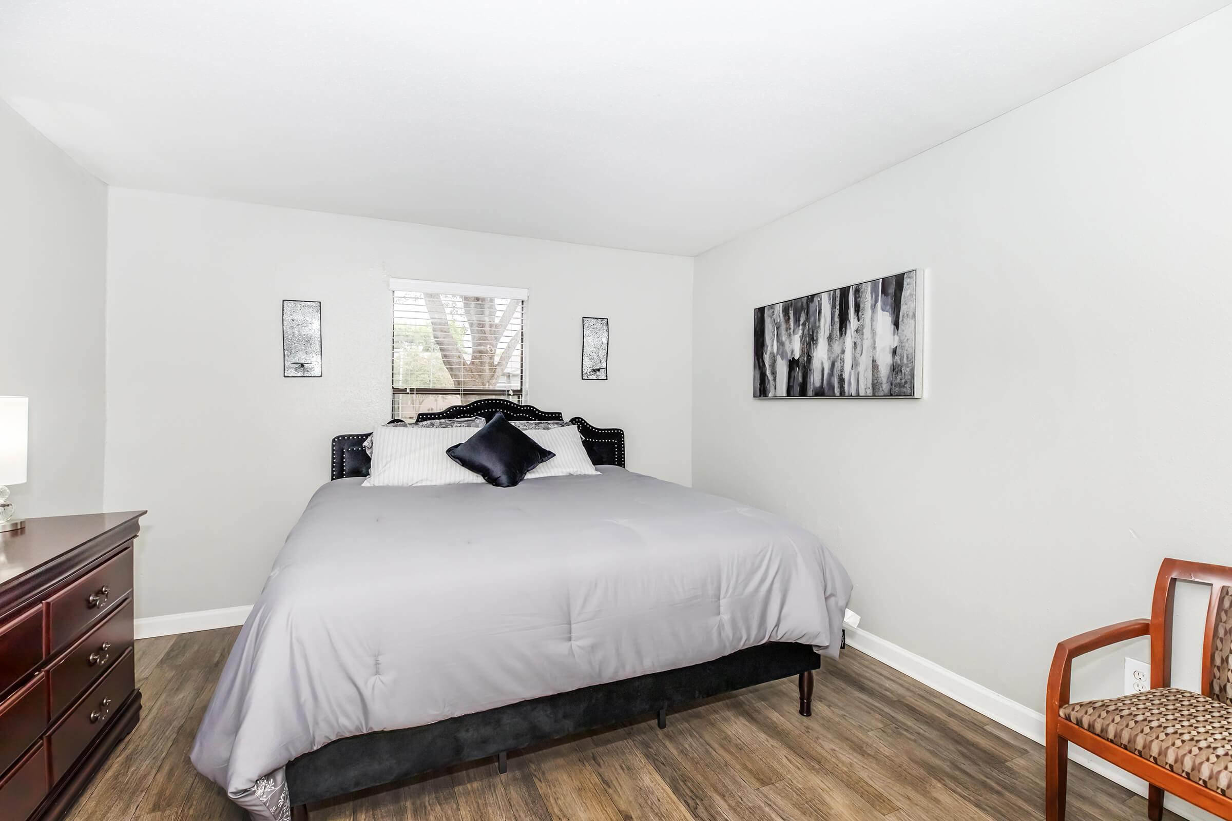 A cozy bedroom featuring a large bed with gray bedding, a dark headboard, and decorative pillows. The walls are painted light gray, and there are two framed art pieces hanging. A window with blinds allows natural light, and a wooden dresser sits against one wall. A chair is positioned in the corner, adding a homey touch.