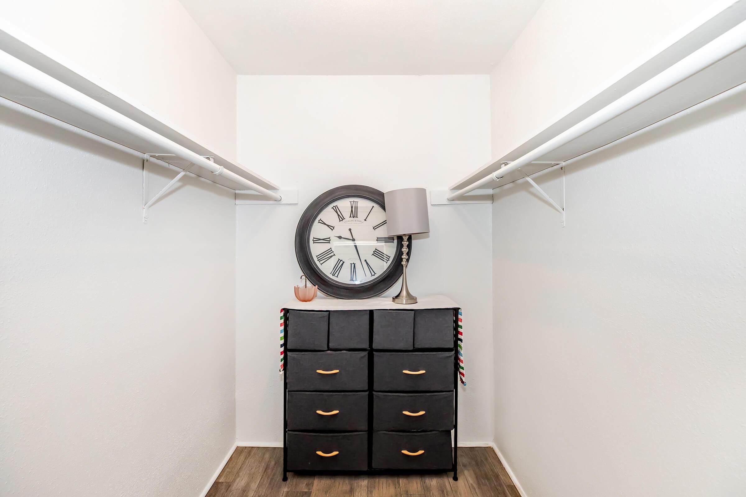 A spacious closet with white walls and two hanging rods. In the center, there is a tall, black dresser with six drawers, topped with a round wall clock displaying Roman numerals and a lamp. The floor is covered in dark wooden planks, and the overall look is clean and organized.