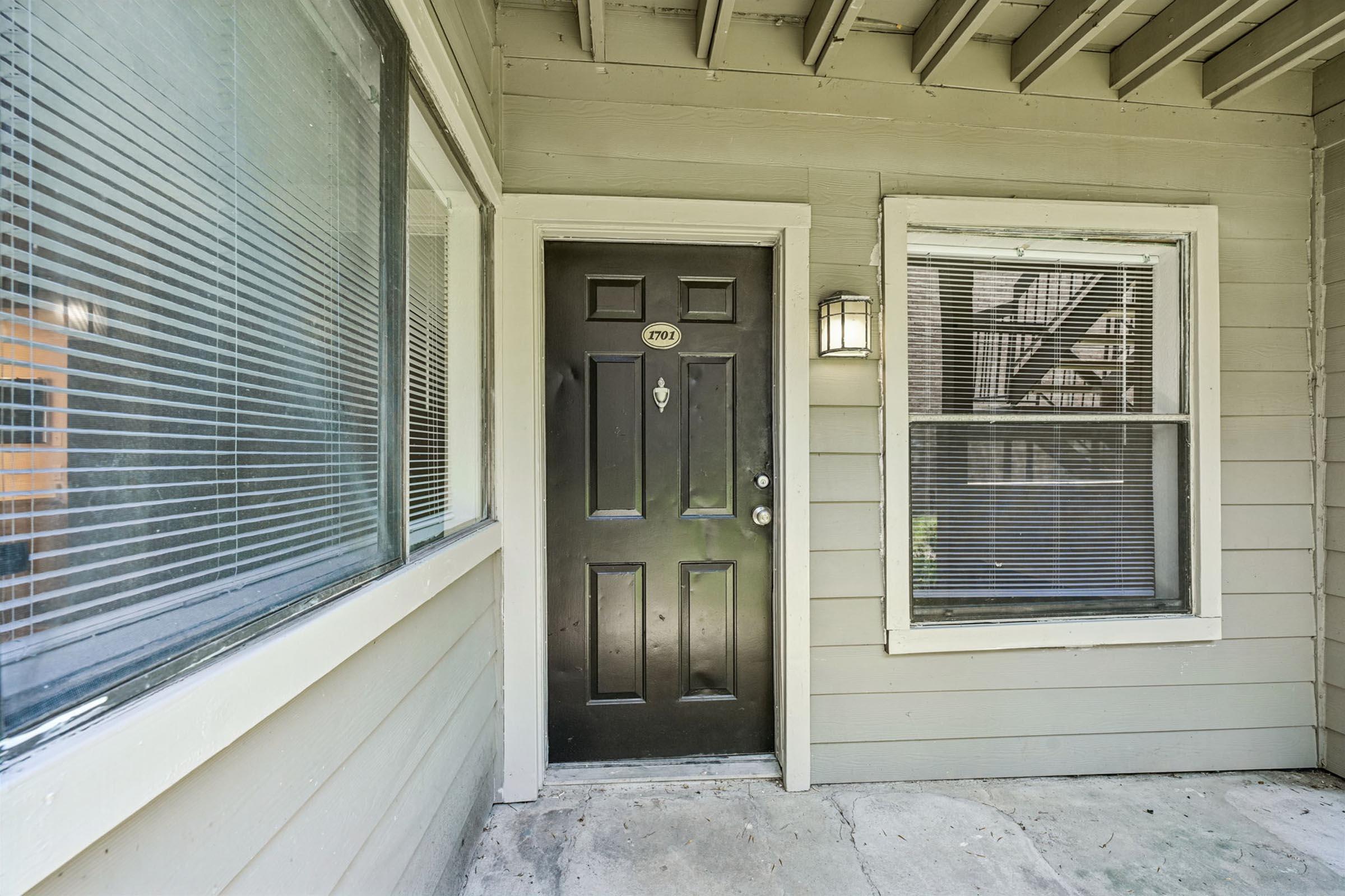A view of a house entrance featuring a black door with the number "203" on it. There are two windows with blinds on either side of the door. The exterior is painted light gray, and the area is partially covered by a roof overhang. The ground shows a concrete surface.